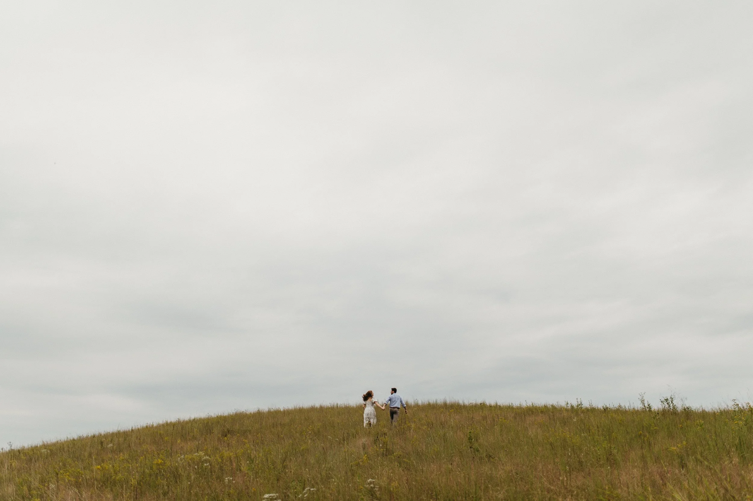 A couple holding hands walking on a grassy hill under a cloudy sky.