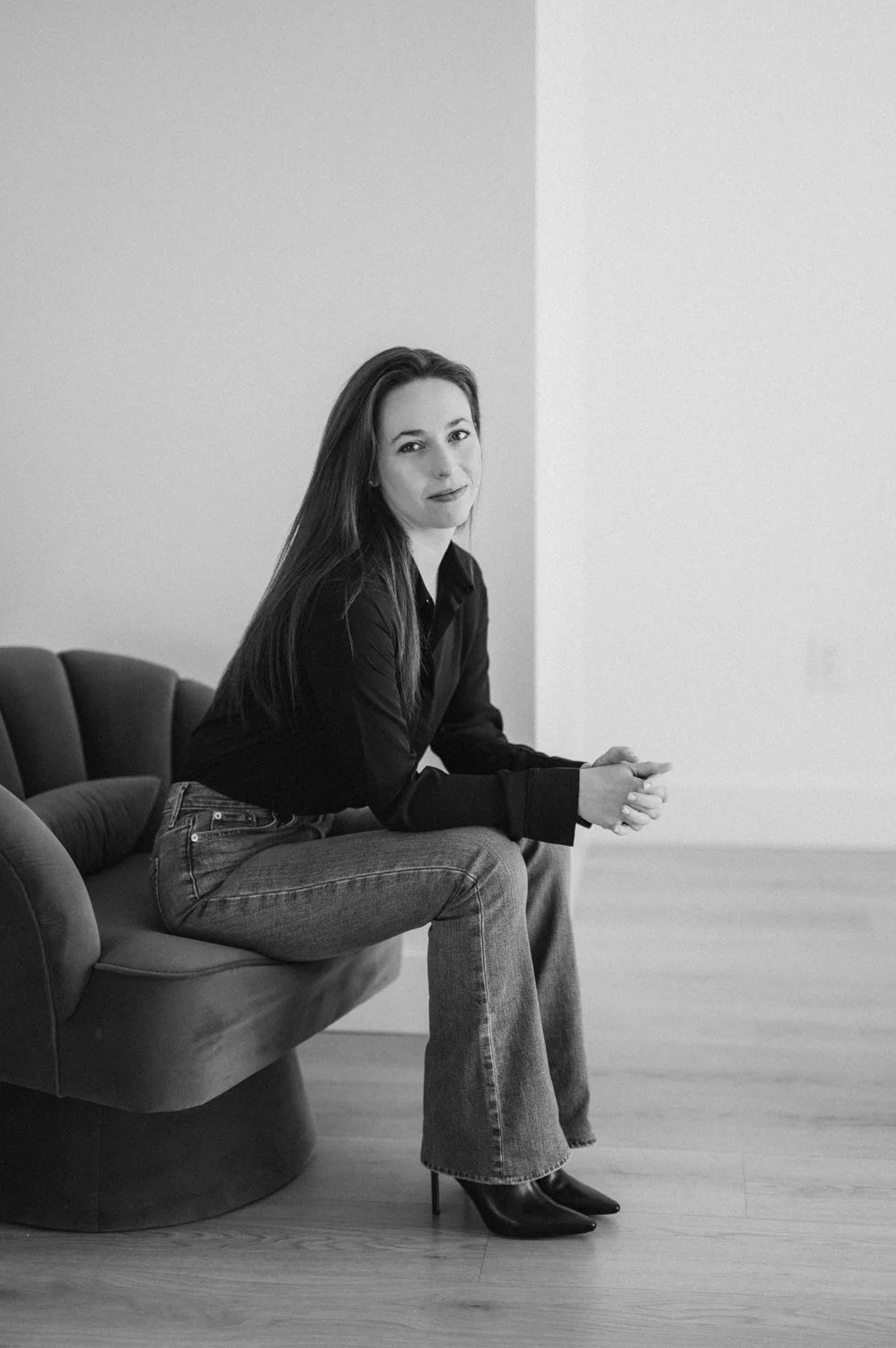 A woman sitting on a modern black sofa in a minimalist room, dressed in a black shirt, jeans, and black high heels, looking at the camera with a slight smile.