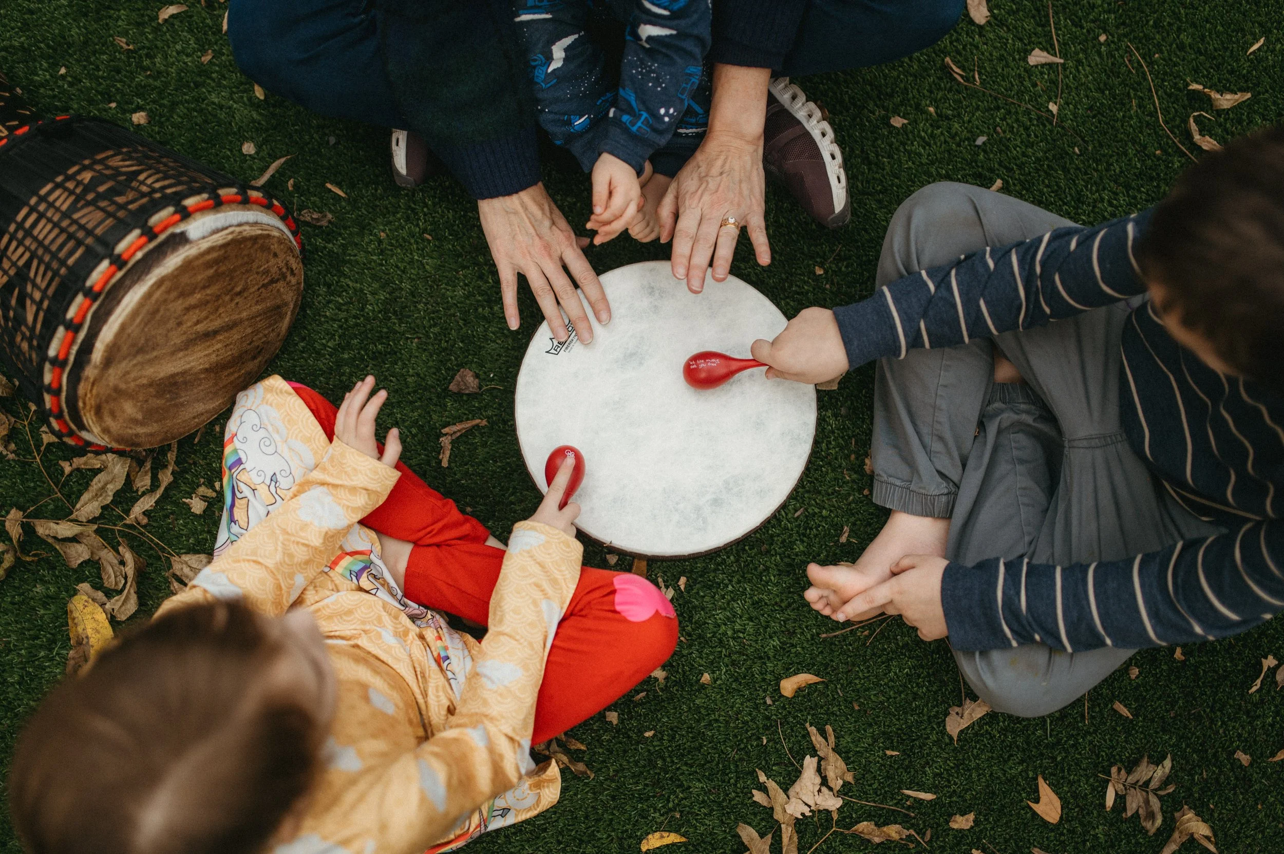 Three children and an adult sitting on grass, playing a game with red maracas and a round drum.
