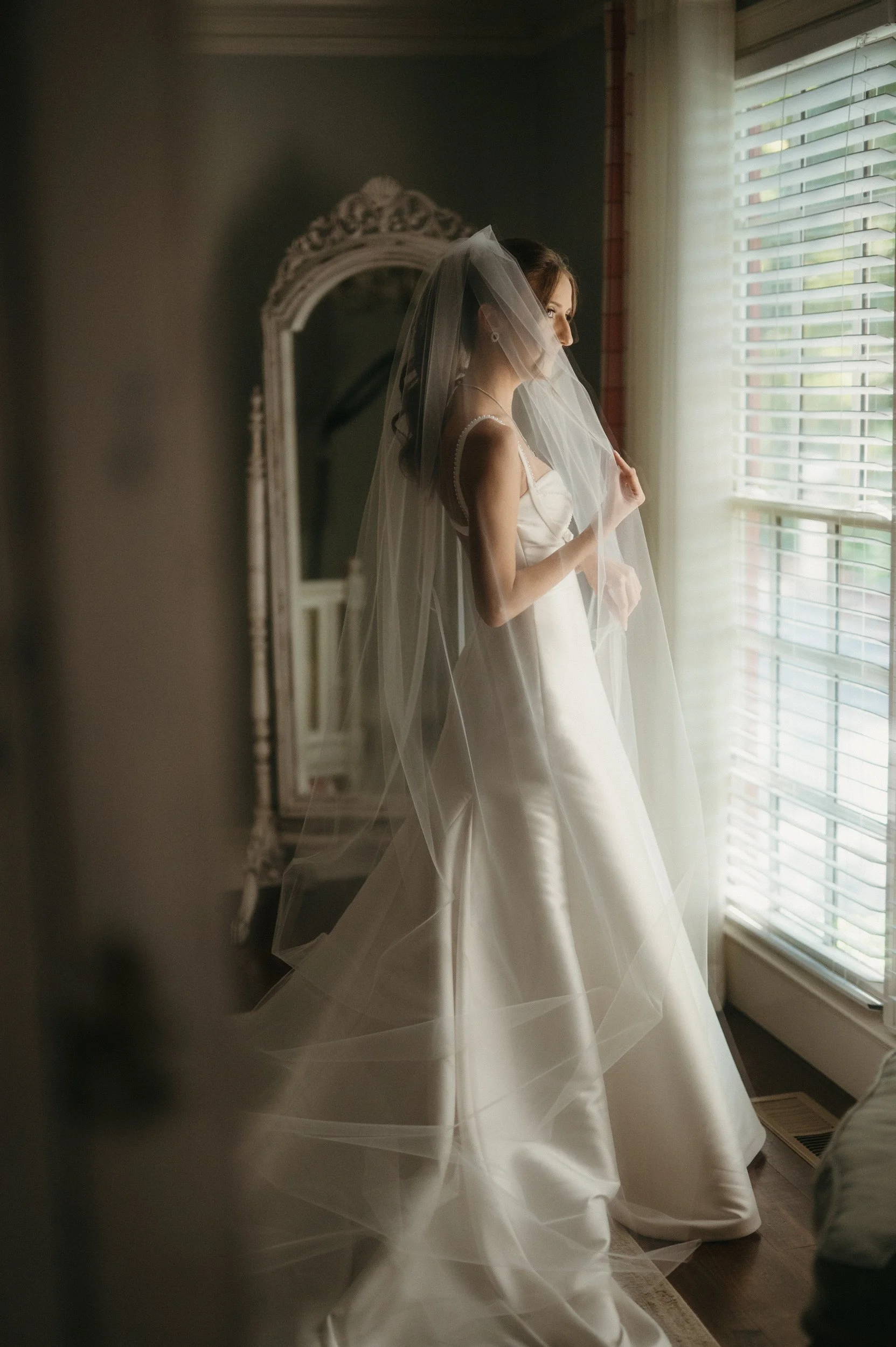 A bride in a white wedding dress and veil standing by a window with blinds, looking out.