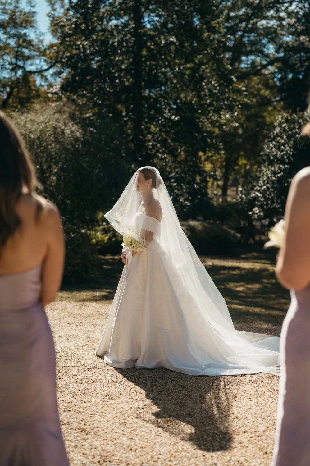 A bride in a white wedding gown with a veil holding a bouquet, standing outdoors during daytime, surrounded by her bridesmaids in lavender dresses.