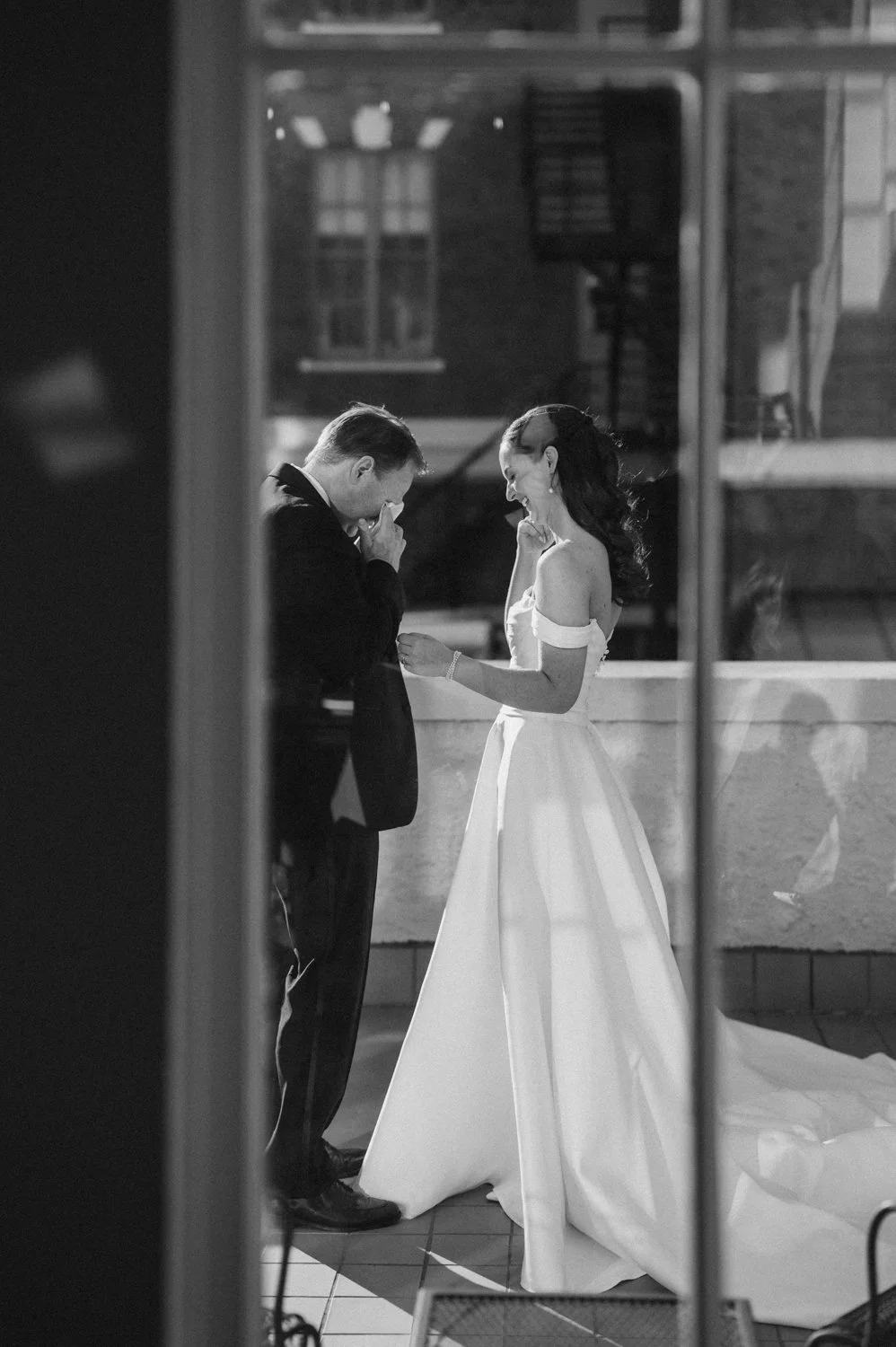 A black-and-white photo of a bride and groom during their wedding ceremony, seen through a window or mirror. The groom is wearing a tuxedo and appears to be emotional, possibly crying, while the bride is smiling and wearing a wedding dress with off-t