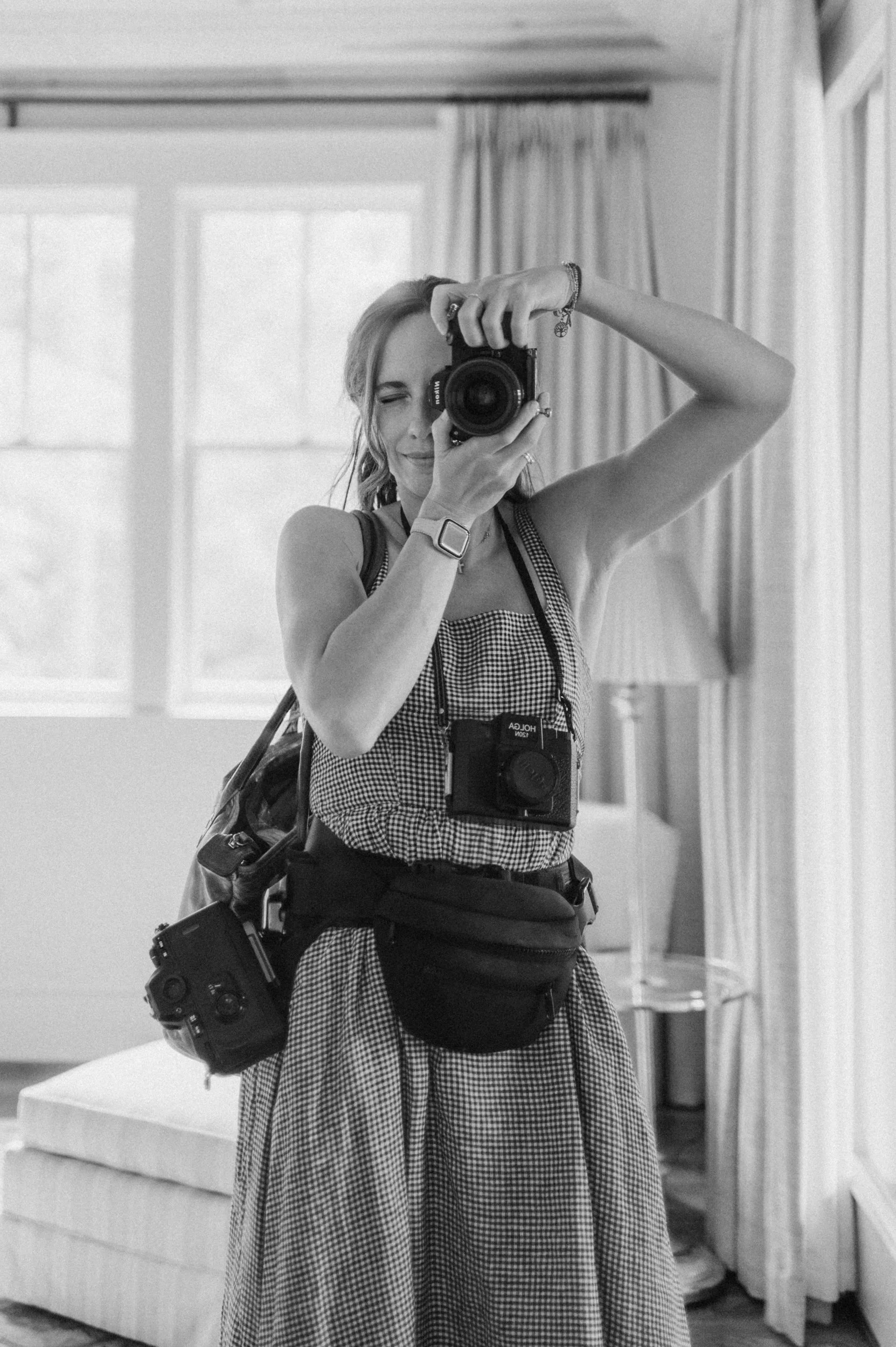 A woman taking a selfie with a camera, in a room with large windows and curtains, wearing a checkered dress, with cameras and gear around her neck and waist.