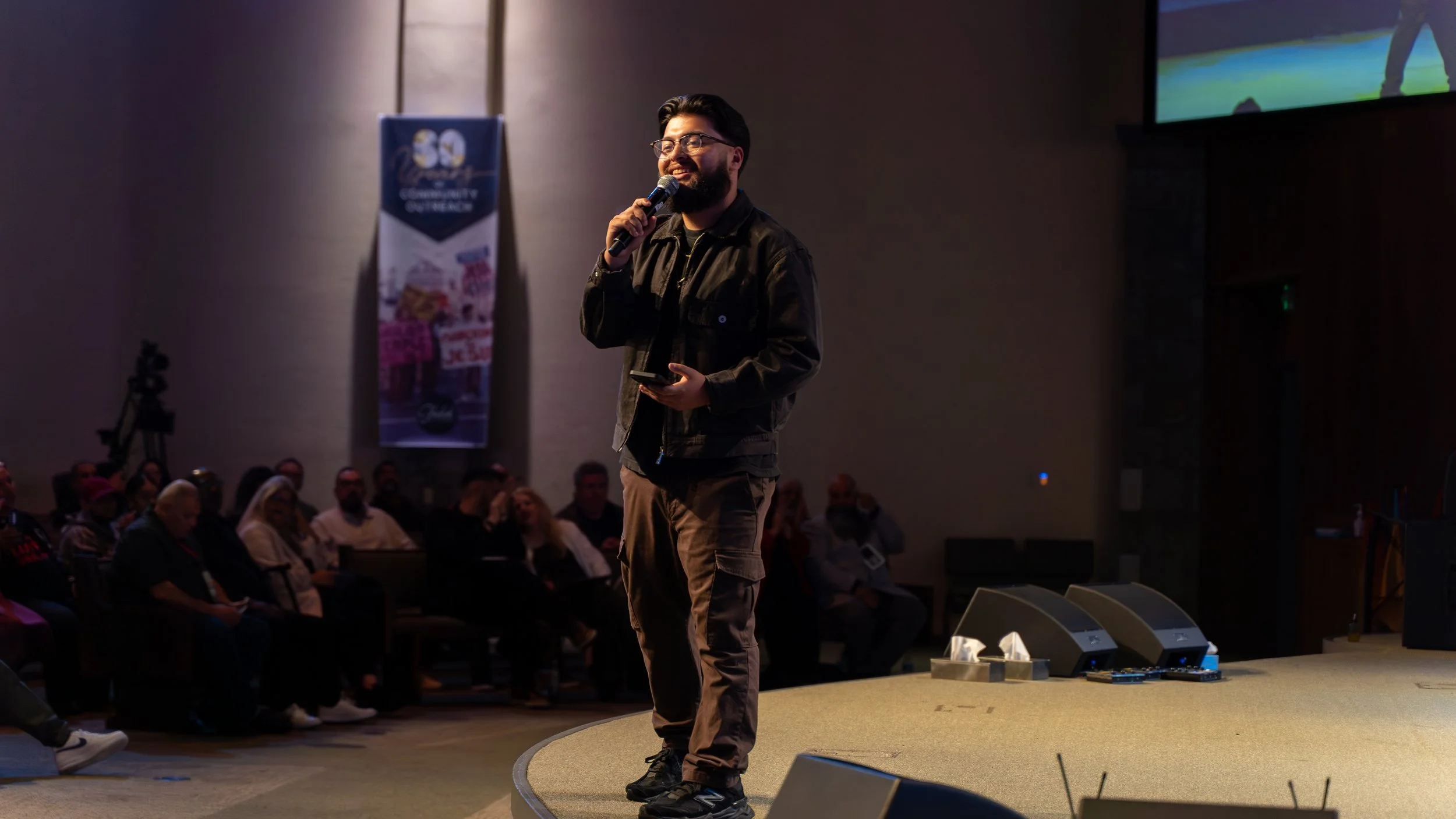 A man with glasses, dark hair, and a beard speaking into a microphone on a stage during a conference. The audience is seated in the background.