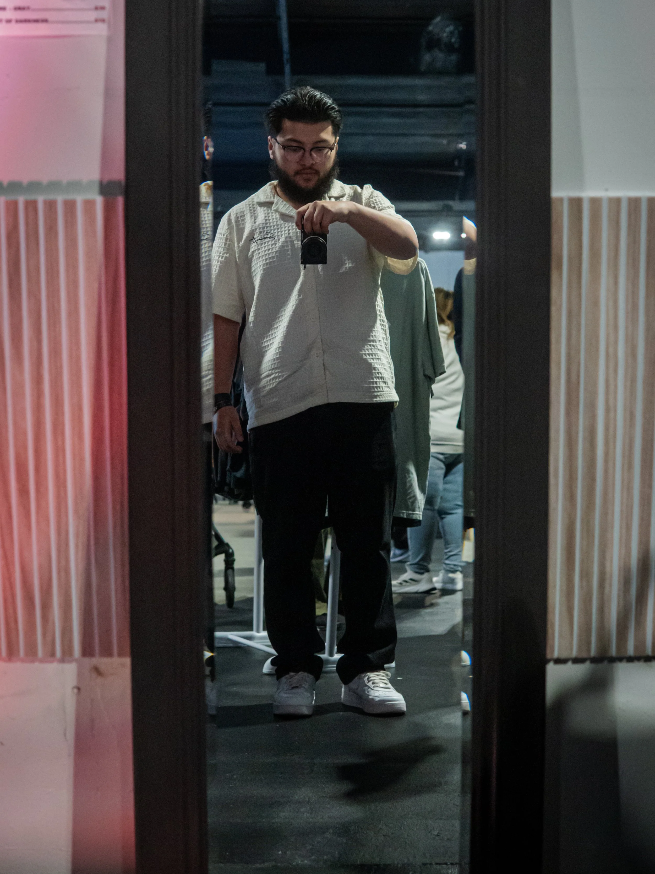 A young man with glasses, a beard, and dark hair takes a selfie in a mirror at a clothing store. He is wearing a white textured short-sleeve shirt, black pants, and white sneakers. There are other shoppers and clothing items visible in the background.