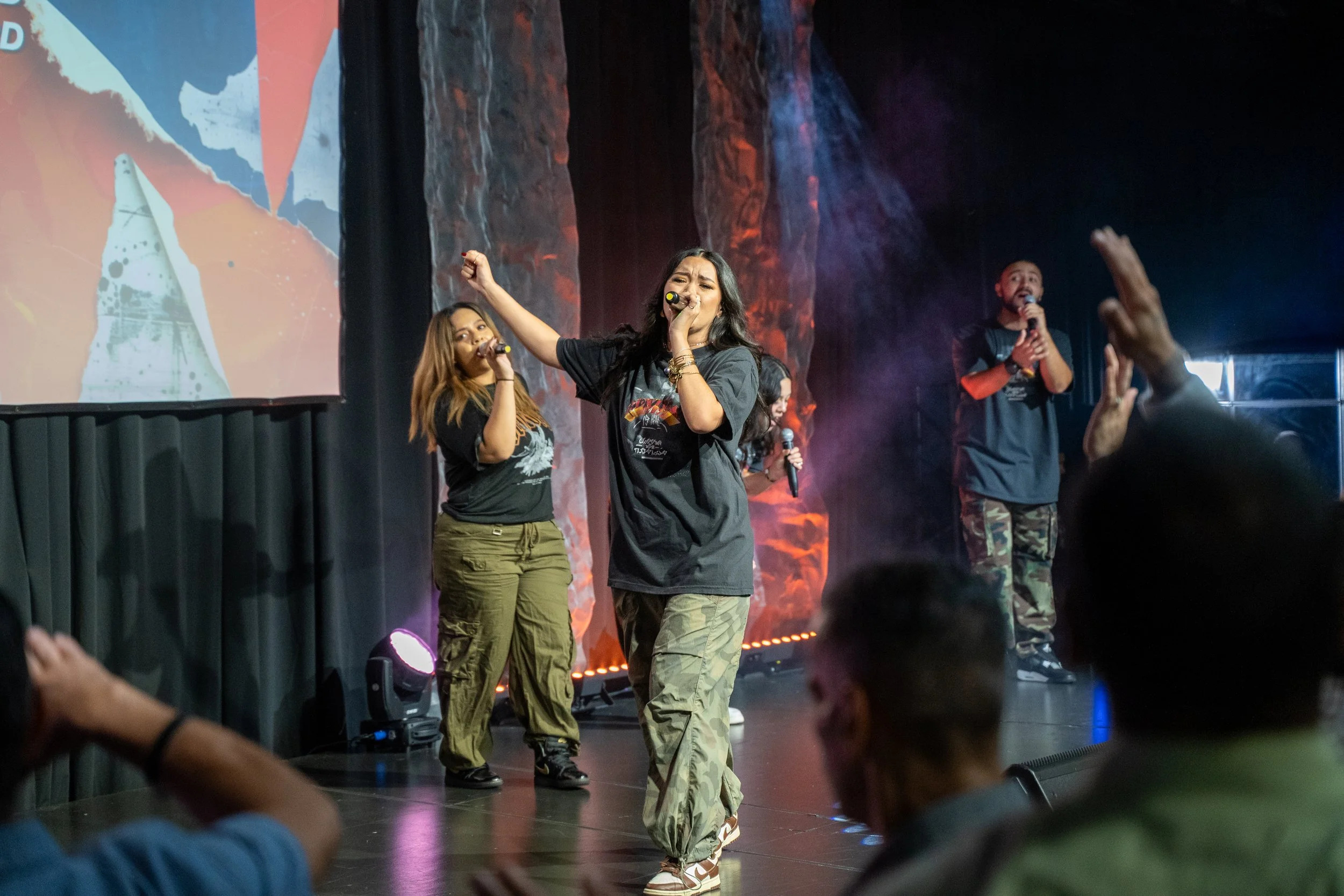 A group of four people singing on stage, holding microphones, with a colorful background and an audience in front.