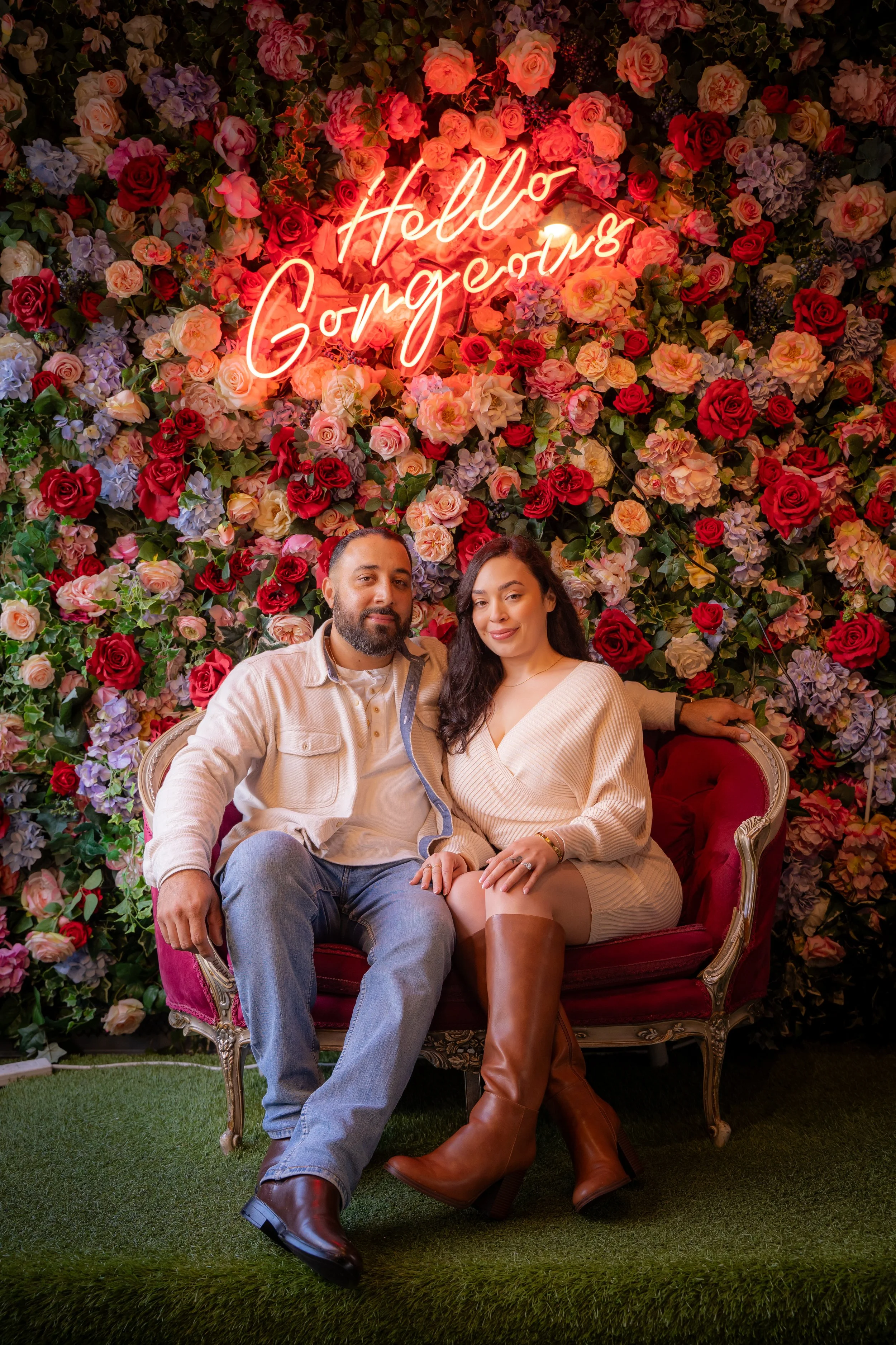 Couple sitting on a red vintage sofa in front of a floral flower wall with a neon sign that says 'Hello Gorgeous'.
