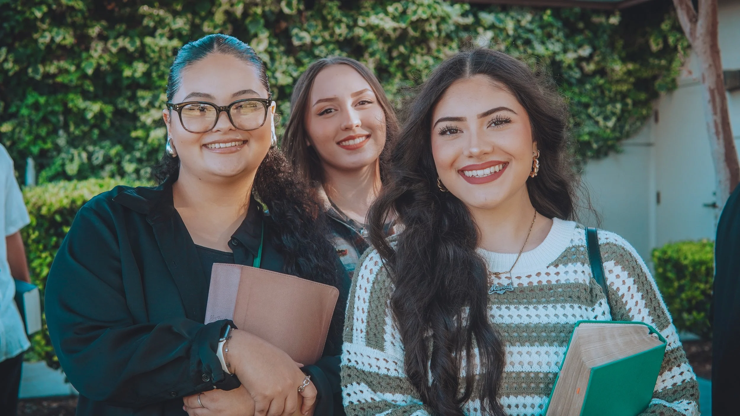 Three young women smiling outdoors, holding books, standing in front of greenery.