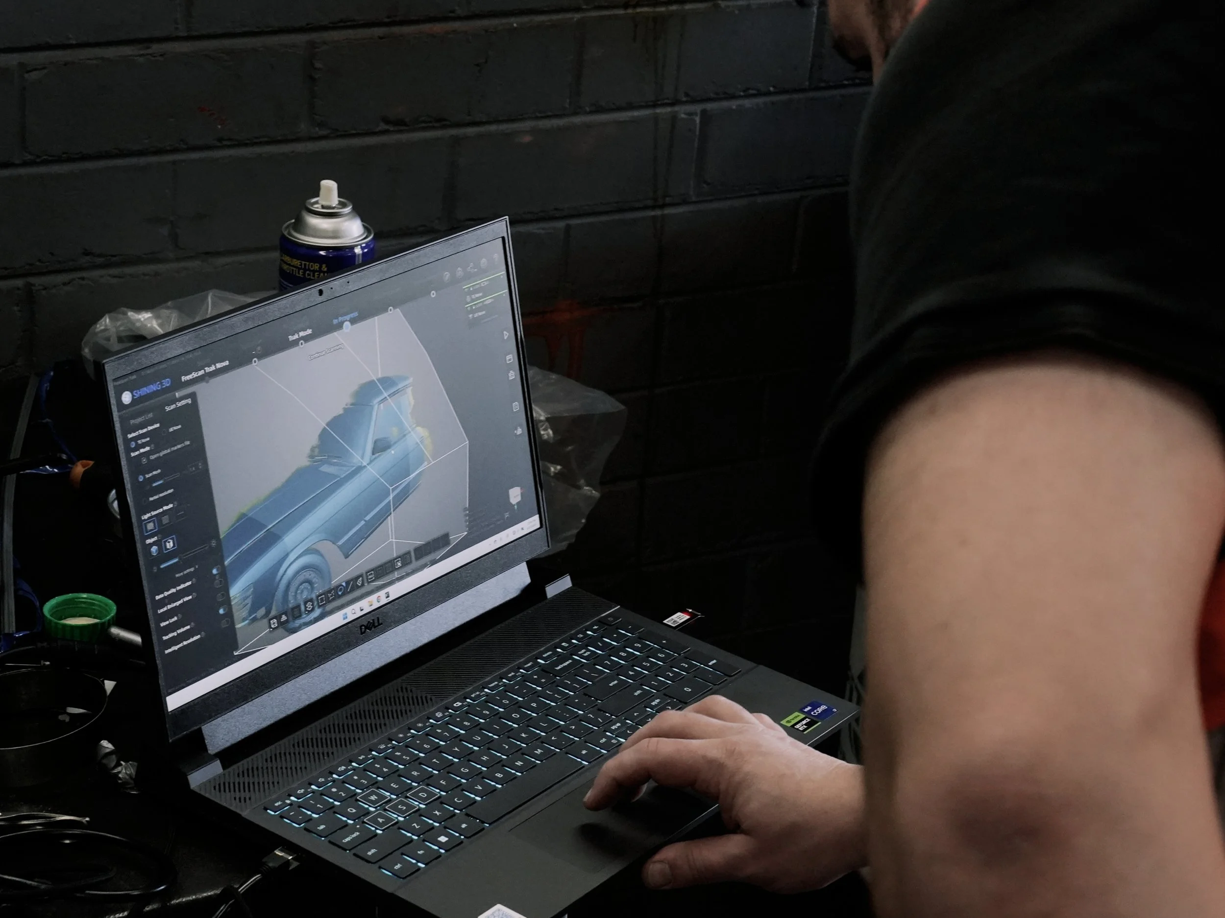 A person working on a Dell laptop displaying 3D car modeling software. The workspace includes a can of compressed air, a plastic bag, and some electronic components on a black brick wall background.