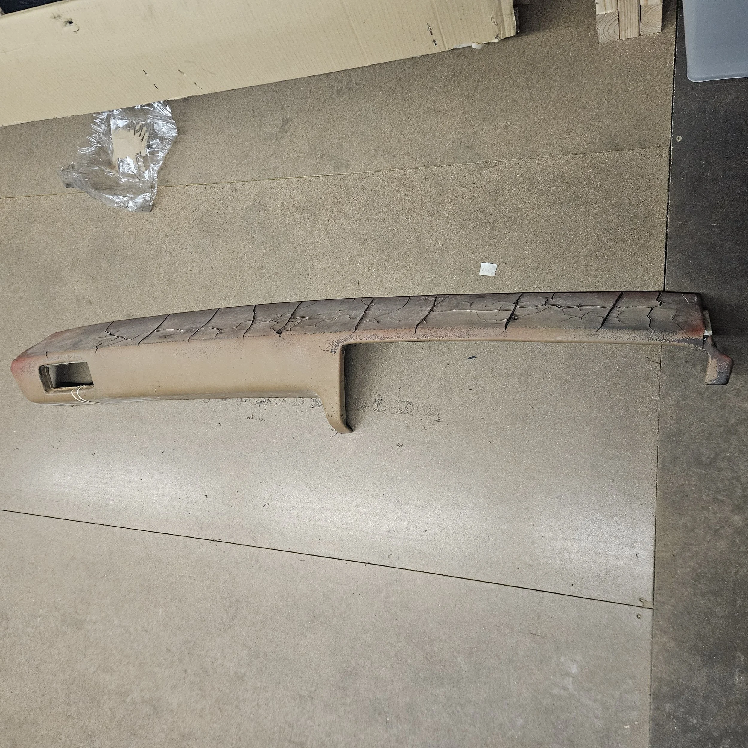 A long, weathered plastic or fiberglass car dashboard with visible cracks and scratches, lying on a tan industrial carpeted floor.