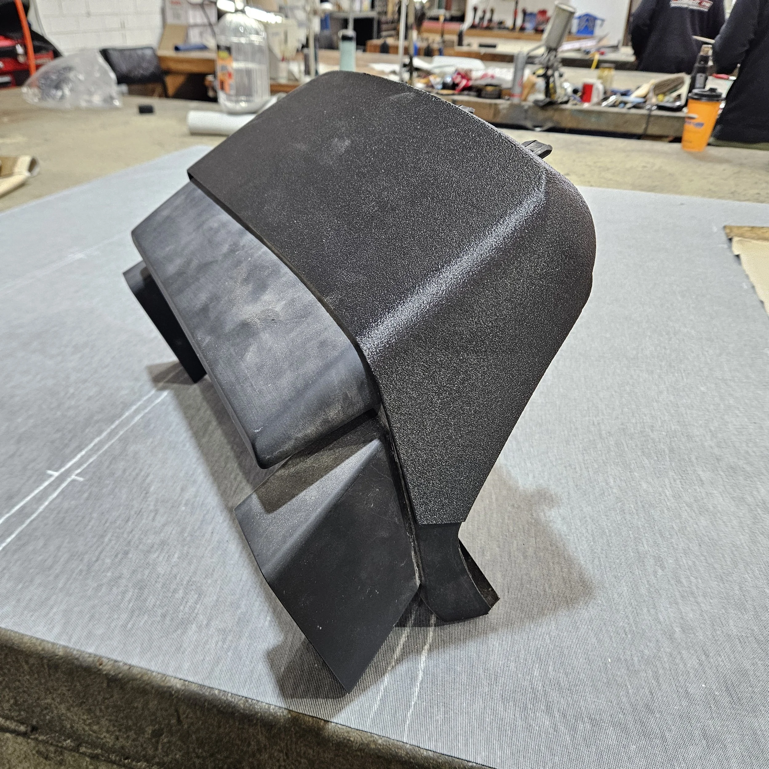 Black mechanical or electronic device with a textured surface, placed on a gray cloth on a workbench, with tools and supplies in the background.