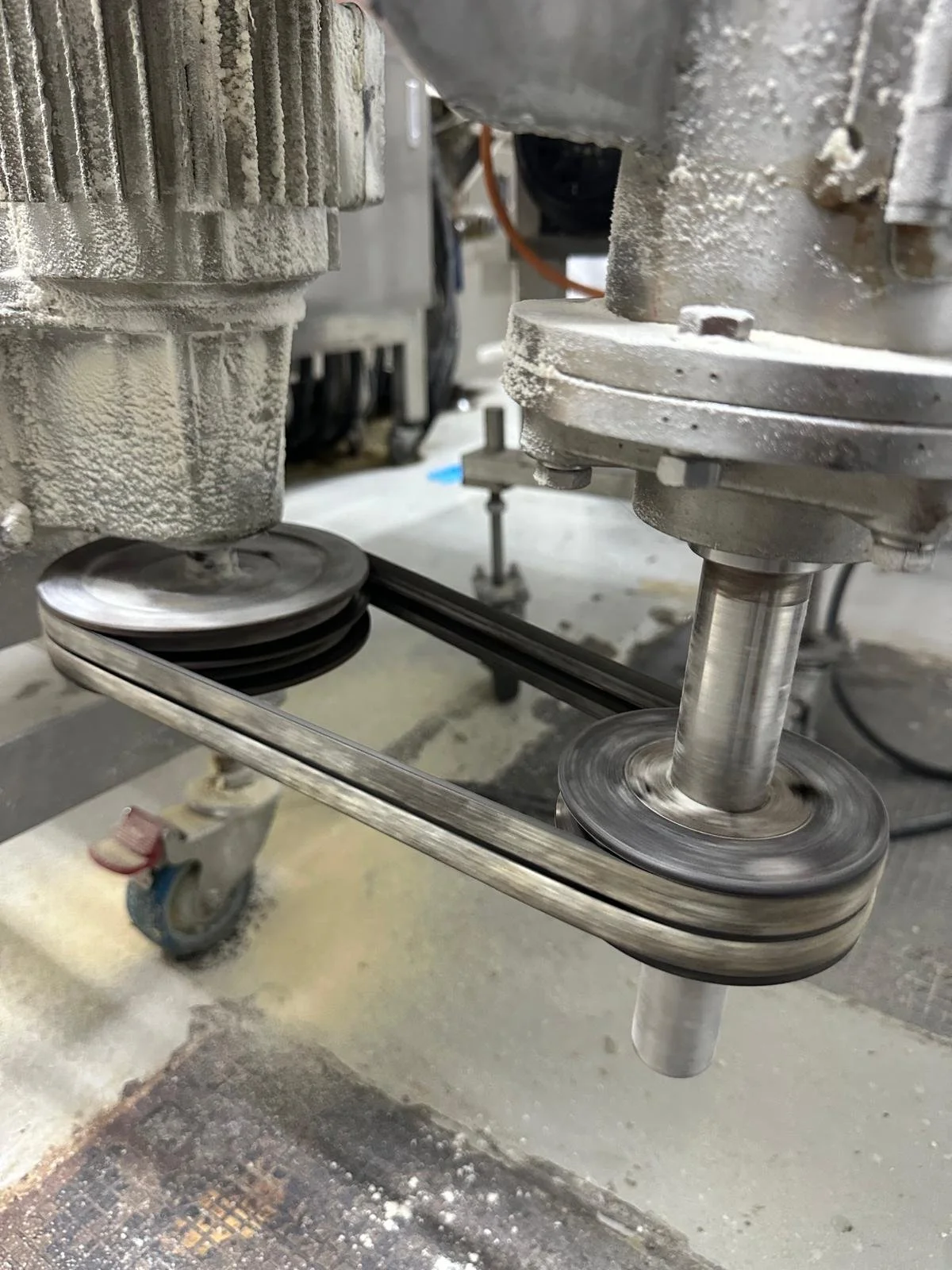 Close-up of a mechanical machine with a belt drive system connecting two pulleys, with a work surface and other industrial equipment in the background.