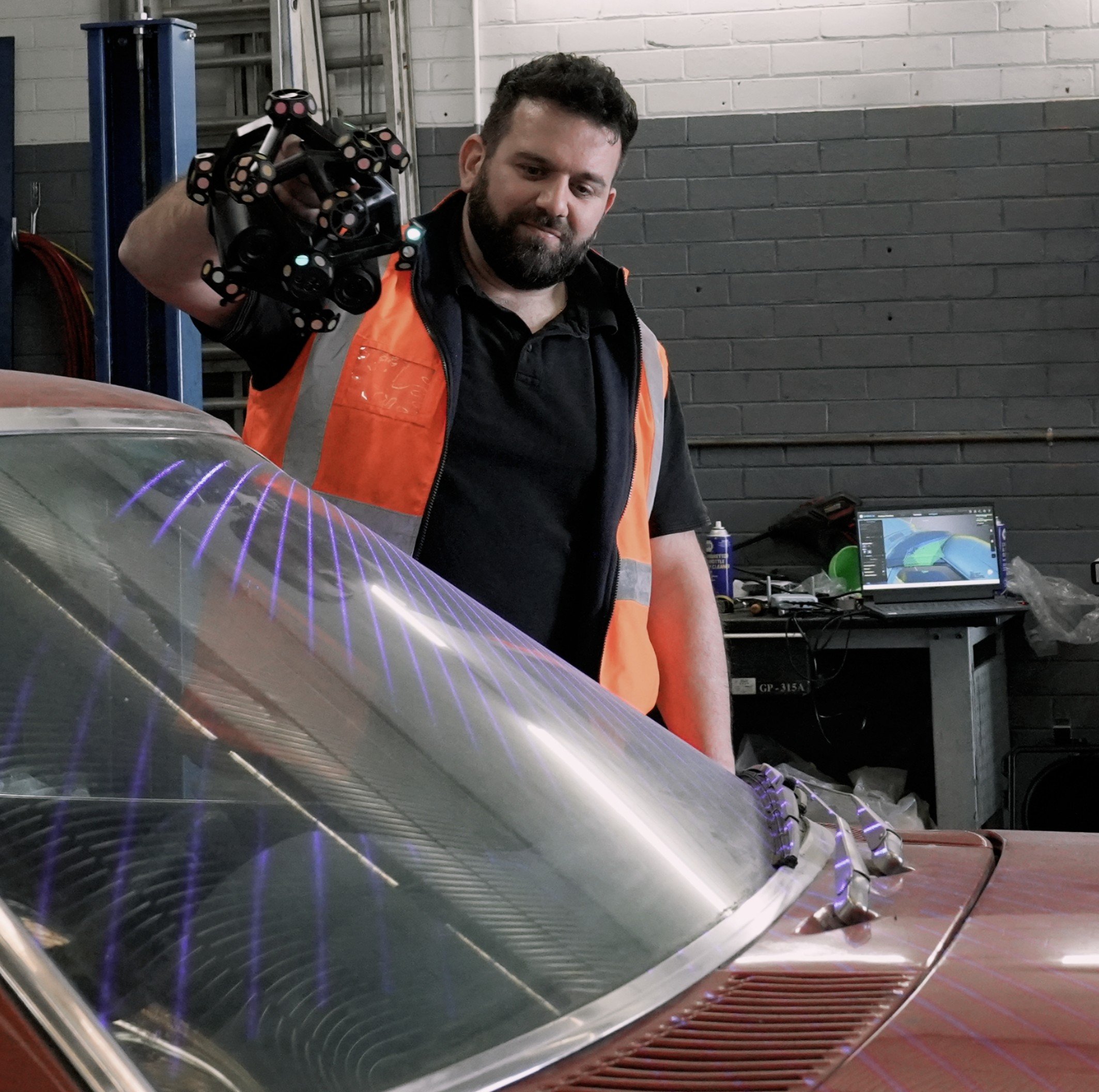 A man wearing an orange safety vest holding a bundle of small black objects with circular patterns, standing next to a vintage car with laser cut patterns on the windshield in a workshop.