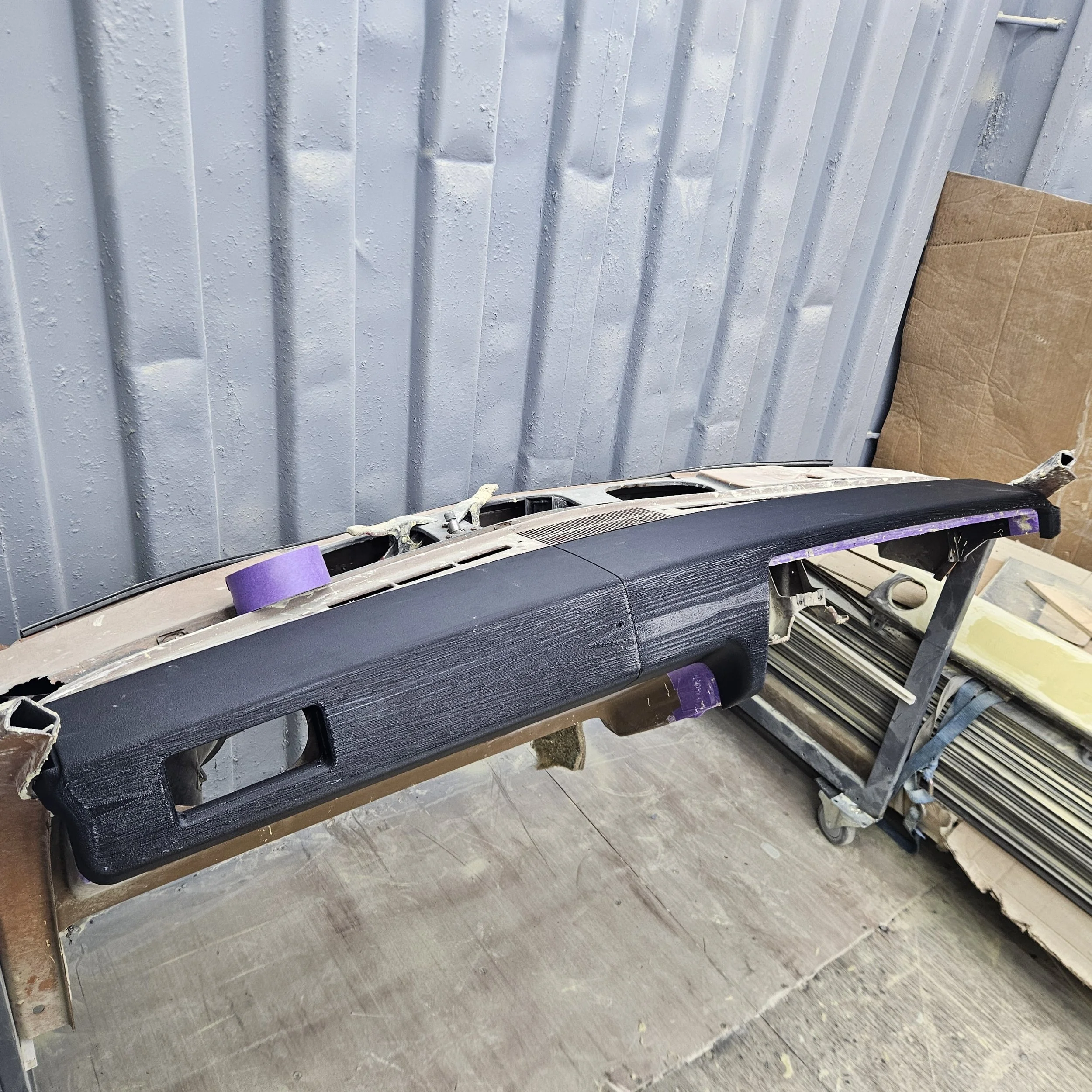 Car dashboard part on a worktable, with purple painter's tape and a graffiti-like black finish, in an industrial workshop.