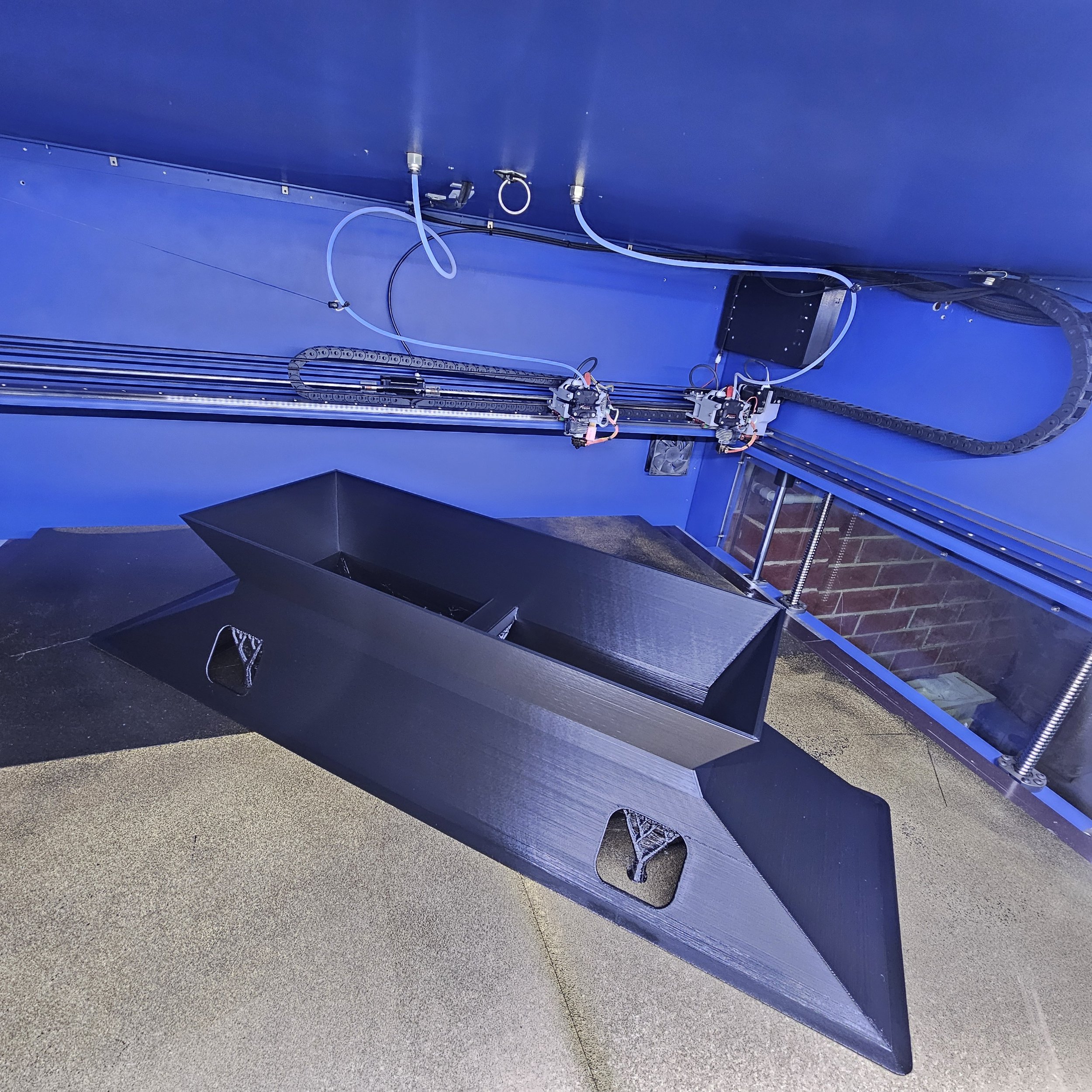 A 3D printer with black parts and a black enclosure, mounted on a blue wall with exposed wires and metal rails.