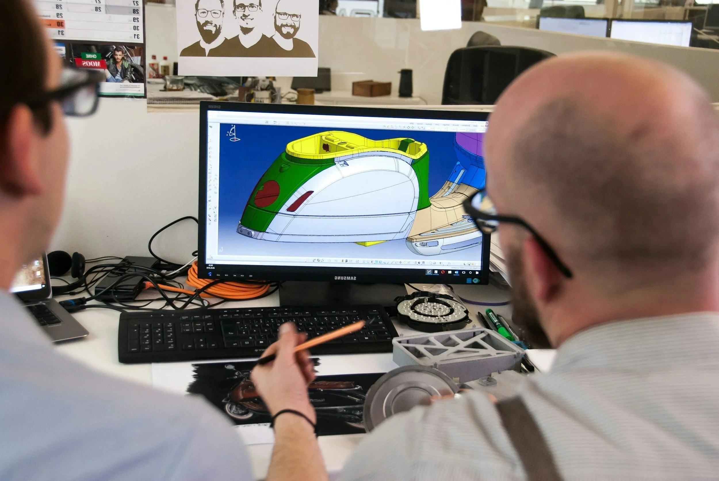 Two designers working together on a computer screen displaying a 3D model of a vehicle, possibly a car or drone, in a busy office environment.