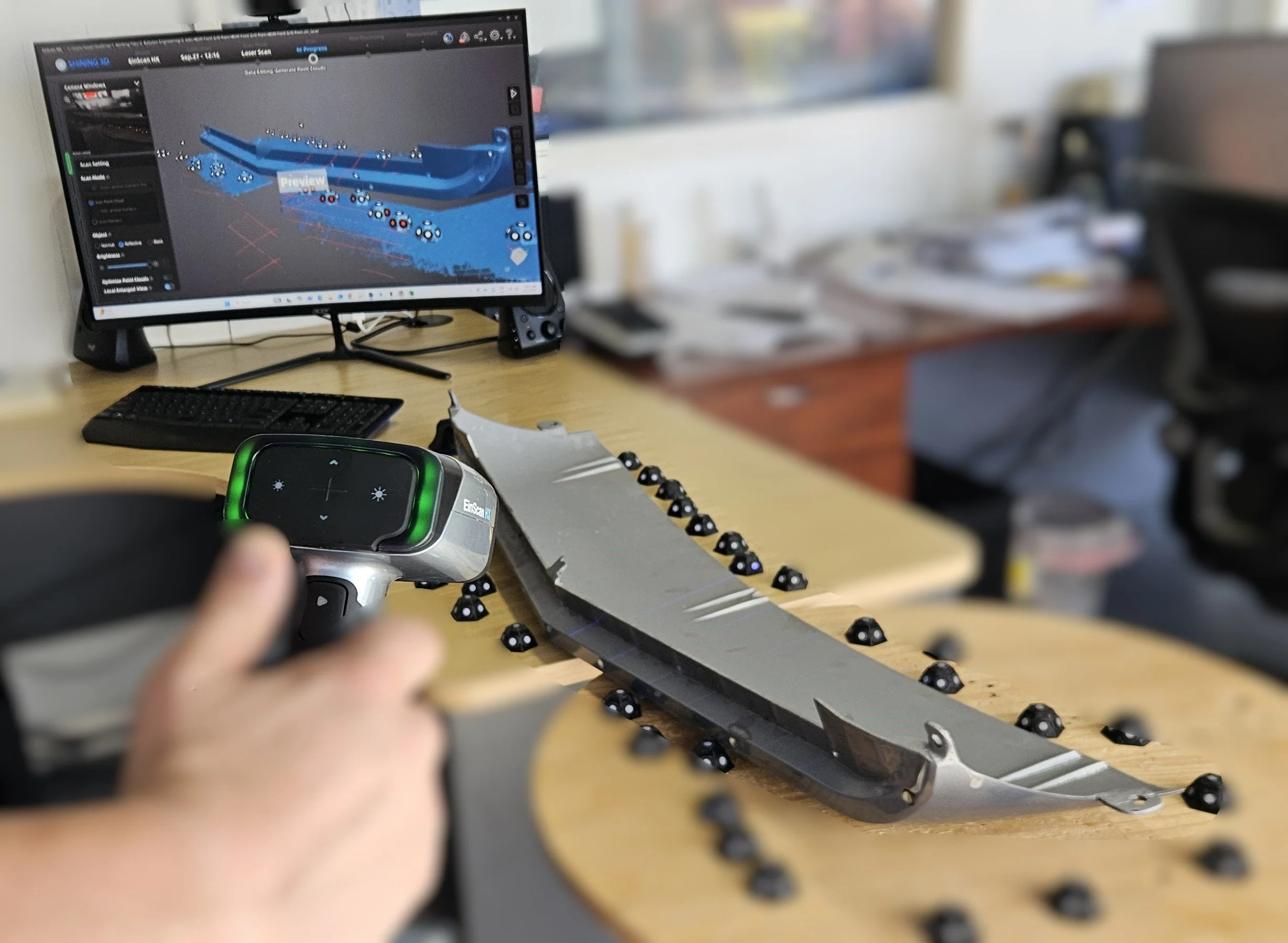 Person using a laser scanner to inspect a car part with plastic black markers, with computer monitor in background showing 3D model of the part.