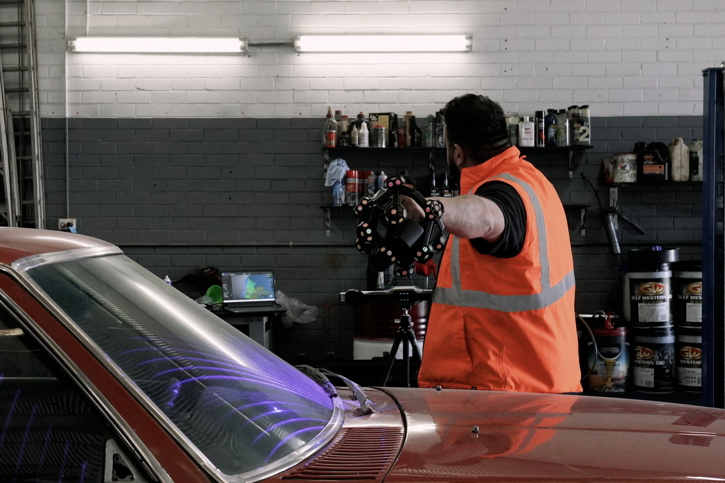 A man wearing an orange safety vest working in an auto repair shop, holding a mechanical part and facing away from the camera, with a classic car in the foreground, a workbench with tools and a laptop in the background.