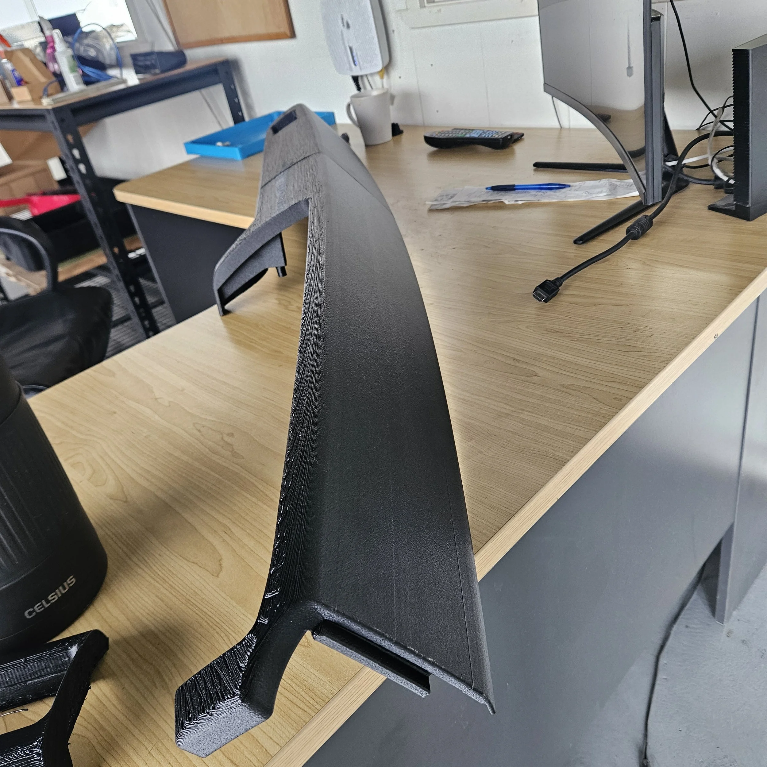 Close-up view of a black 3D-printed object with a curved edge placed on a wooden desk in an office or workshop setting.