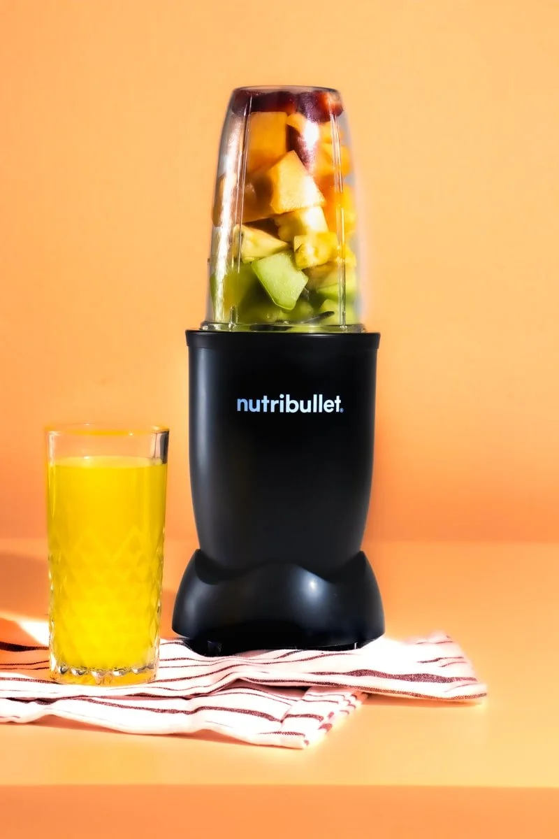 nutri bullet with layered fruit on orange background, orange juice, and white stripped hand towel