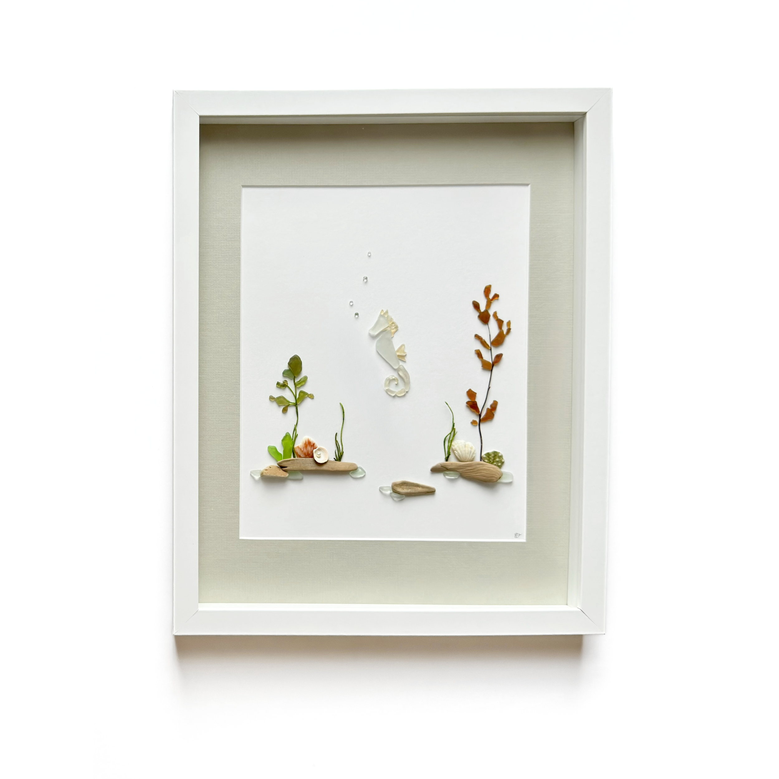 Framed white sea glass seahorse artwork surrounded by seaweed, shells, driftwood, and beach stones.