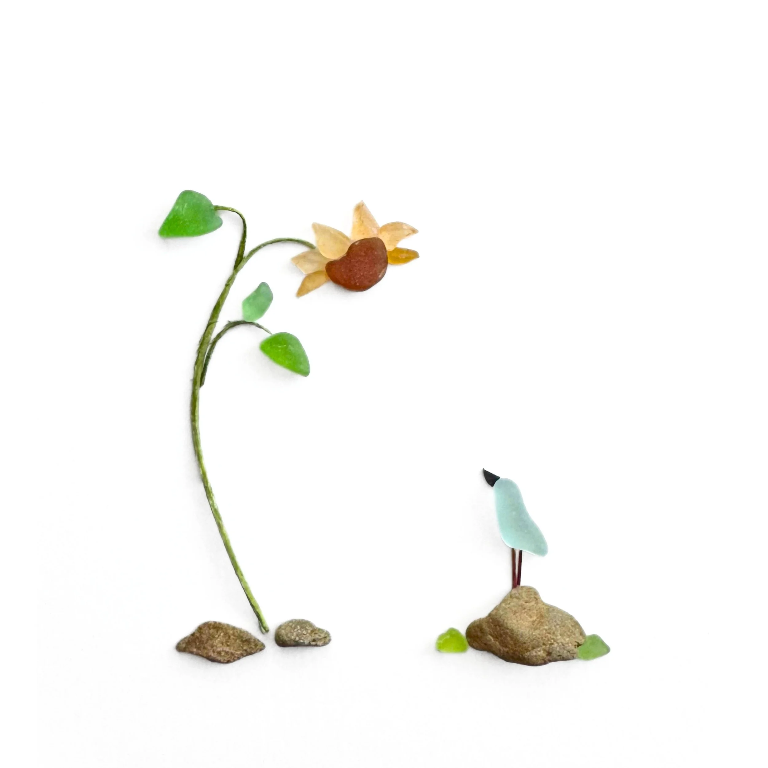 Original sea glass artwork featuring a tiny pale blue bird perched on a beach stone, looking up at a golden sunflower with green sea glass leaves and a curved botanical stem against a white background