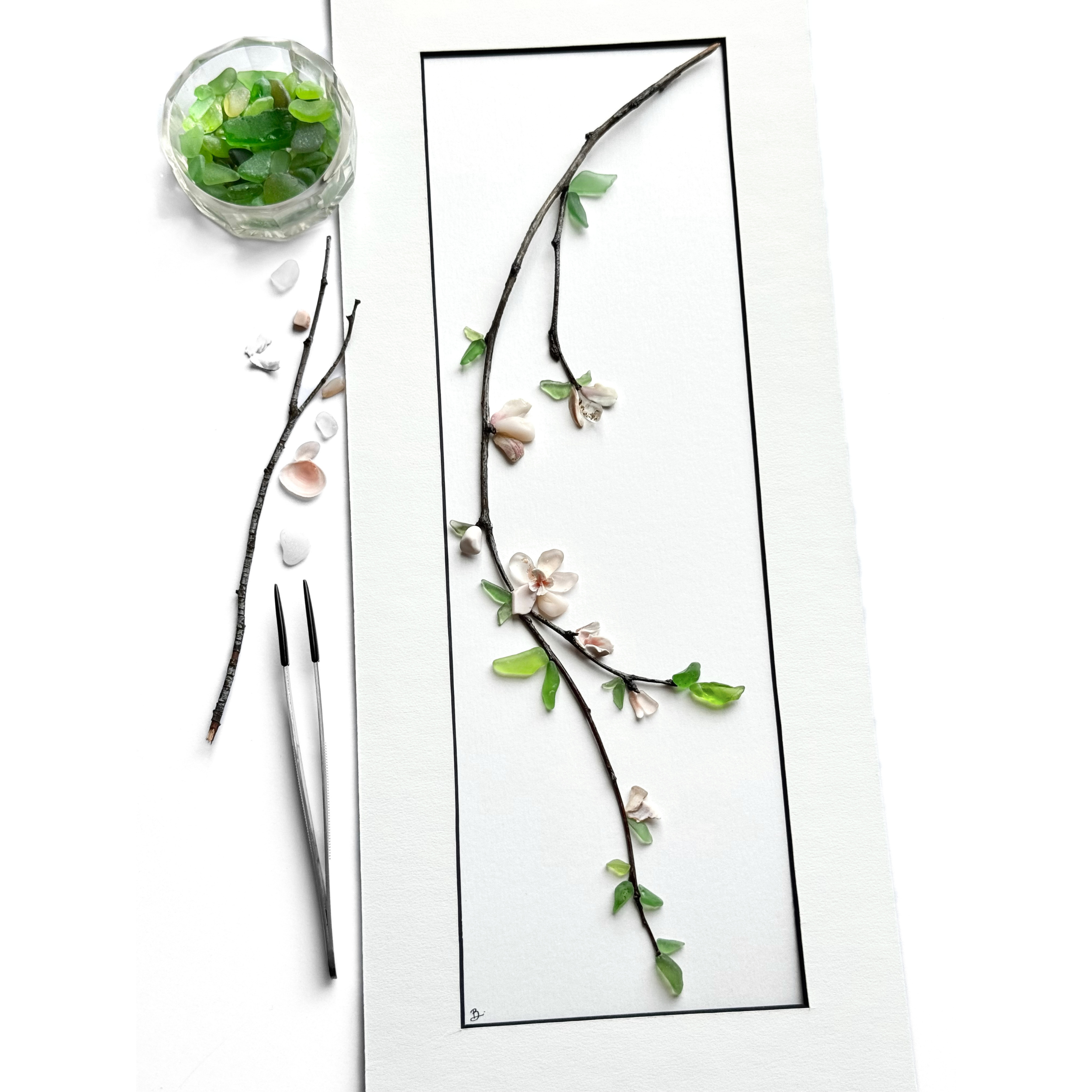 Studio process view of a cherry blossom branch arranged inside a narrow black mat with tweezers, pink shells and green sea glass nearby.