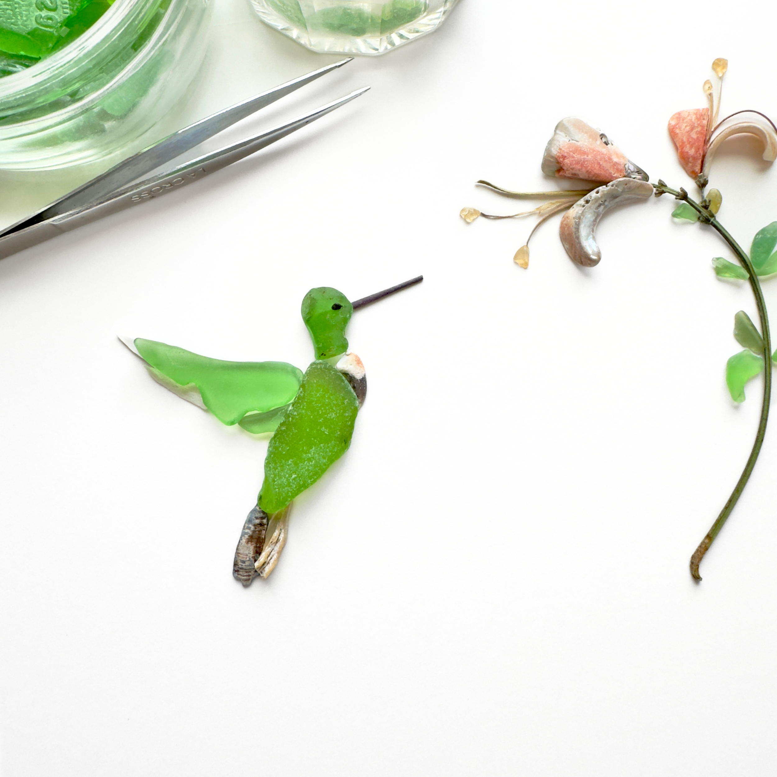 A behind-the-scenes view of a hummingbird approaching soft pink blossoms, with an artist’s tweezers nearby.