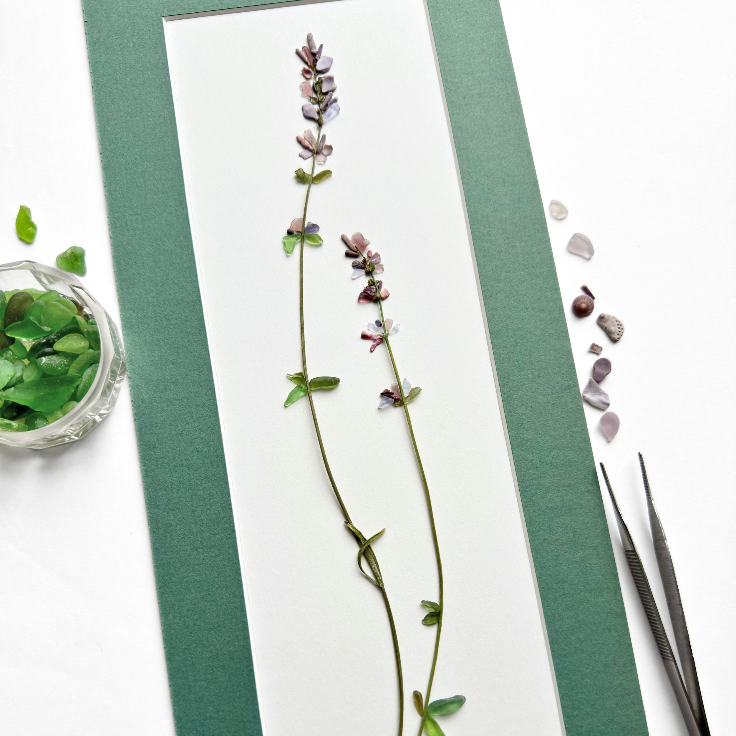 Studio process view of a tall sea glass lavender stem with loose materials and tweezers on a green matted work surface.