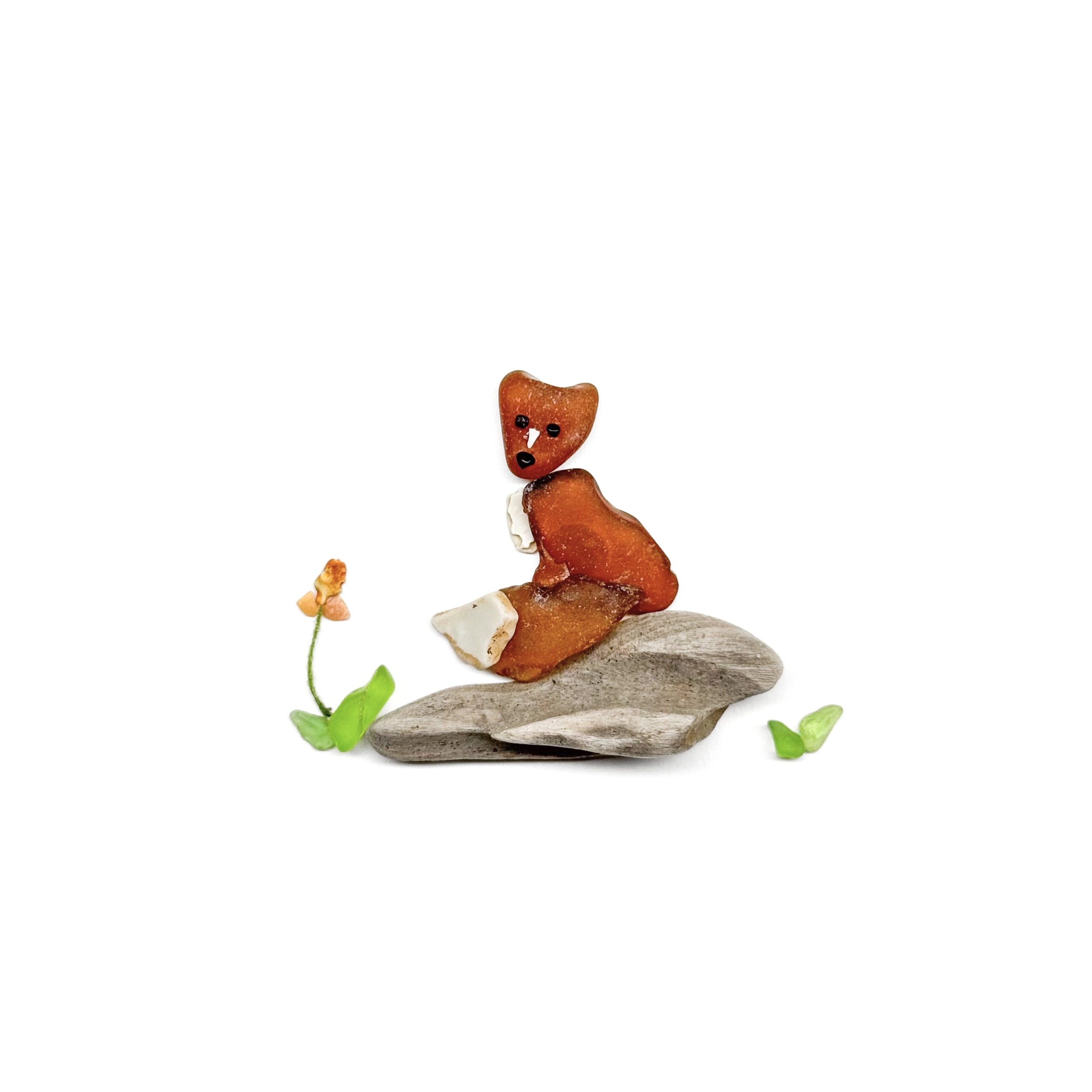 A little sea glass fox sits thoughtfully on driftwood beside a tiny woodland flower.