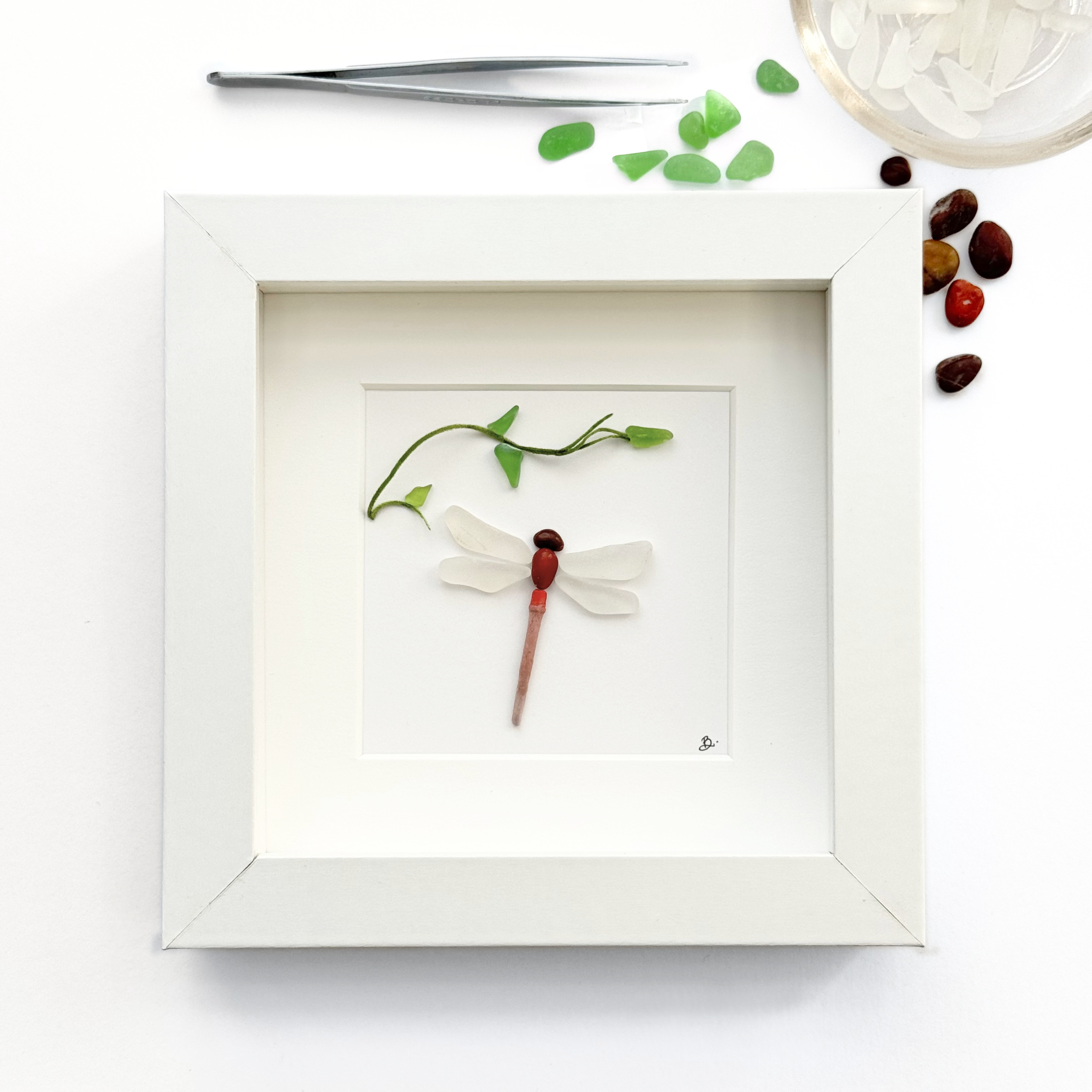 A framed dragonfly created from sea glass, surrounded by a glimpse of the materials that inspired it.