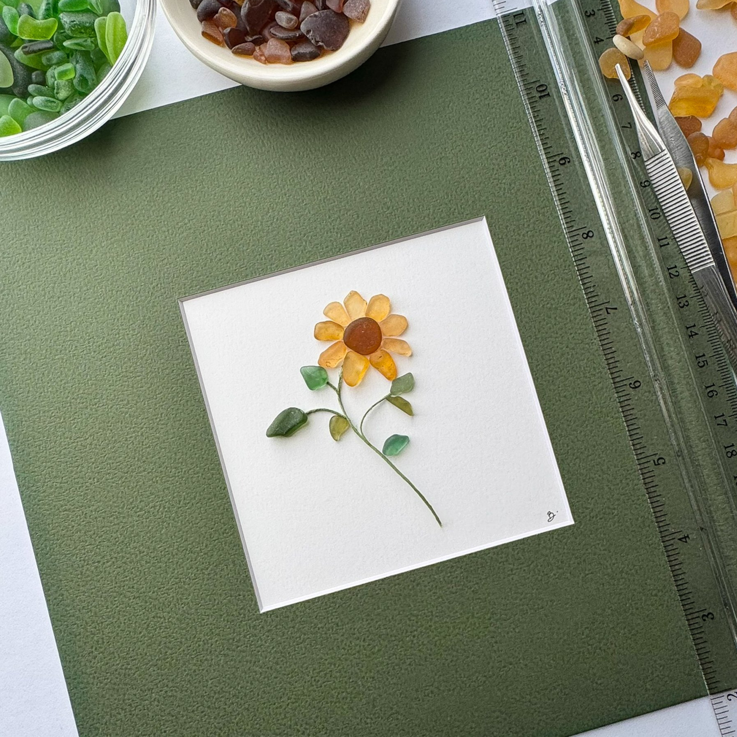 Studio process view of a sea glass sunflower on a green matted work surface with tools and materials nearby.