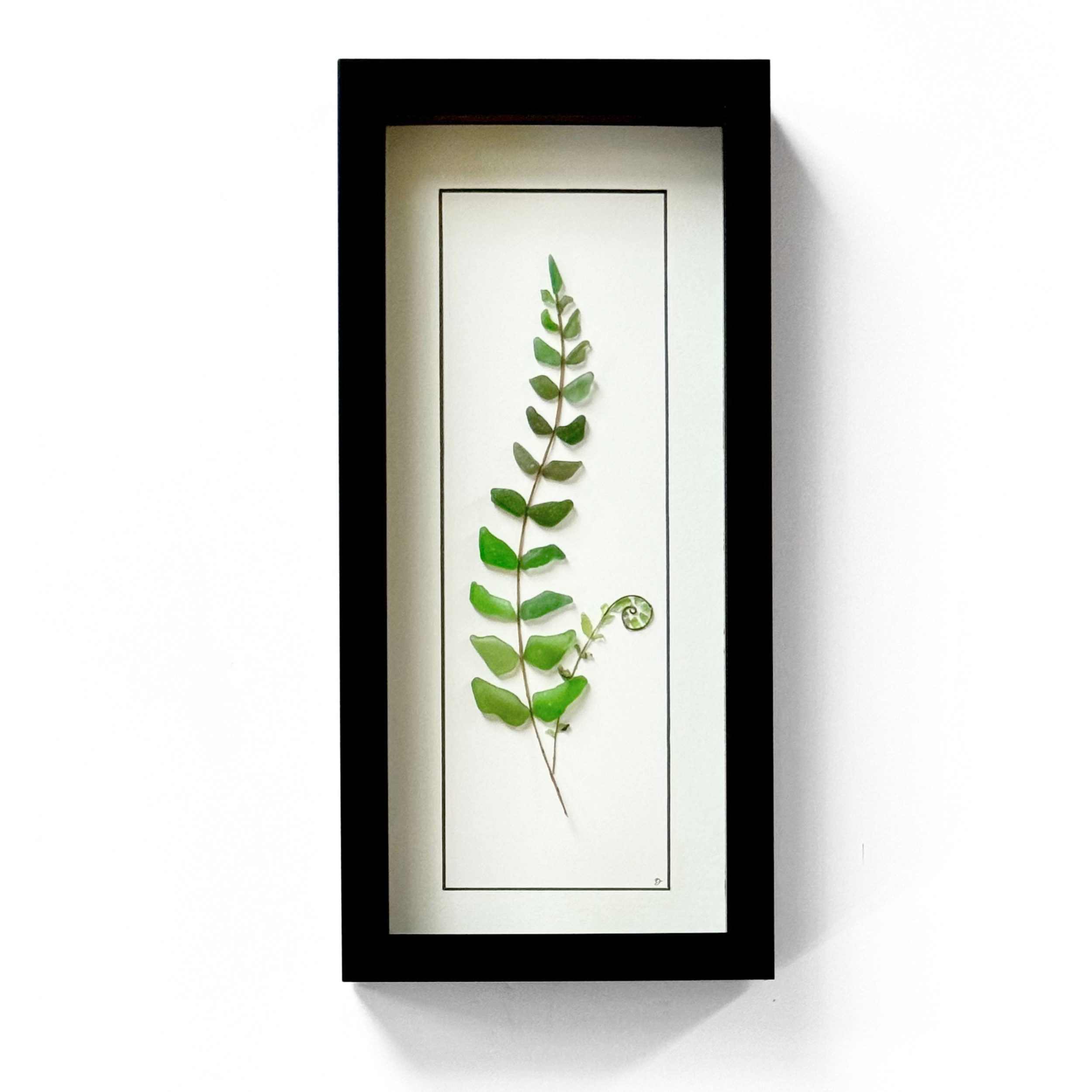 A framed forest fern created from carefully arranged green sea glass, with a tiny spiral fiddlehead tucked beside its stem.