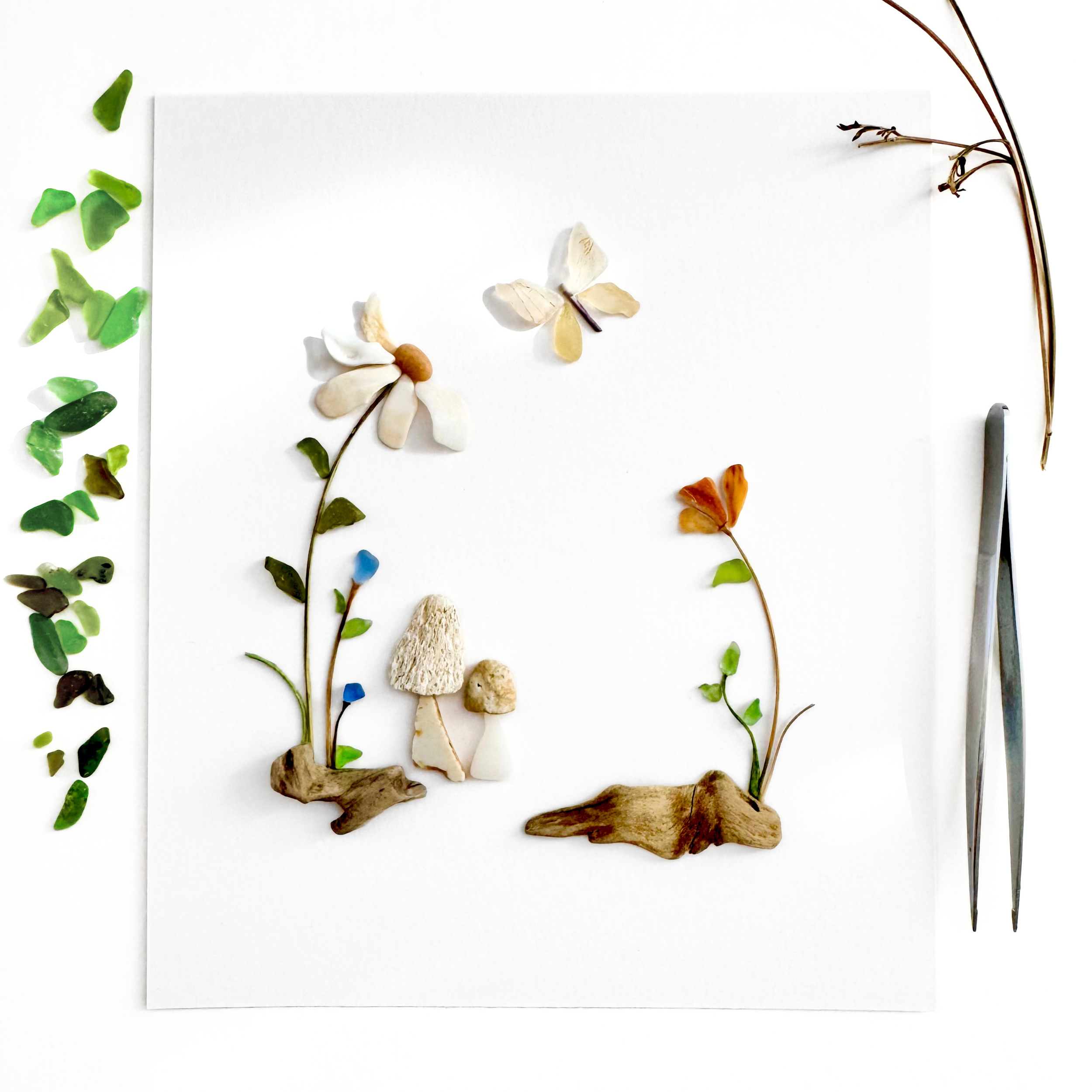 Studio process scene with sea glass wildflowers, butterflies, trailing vines, natural materials, and tweezers.