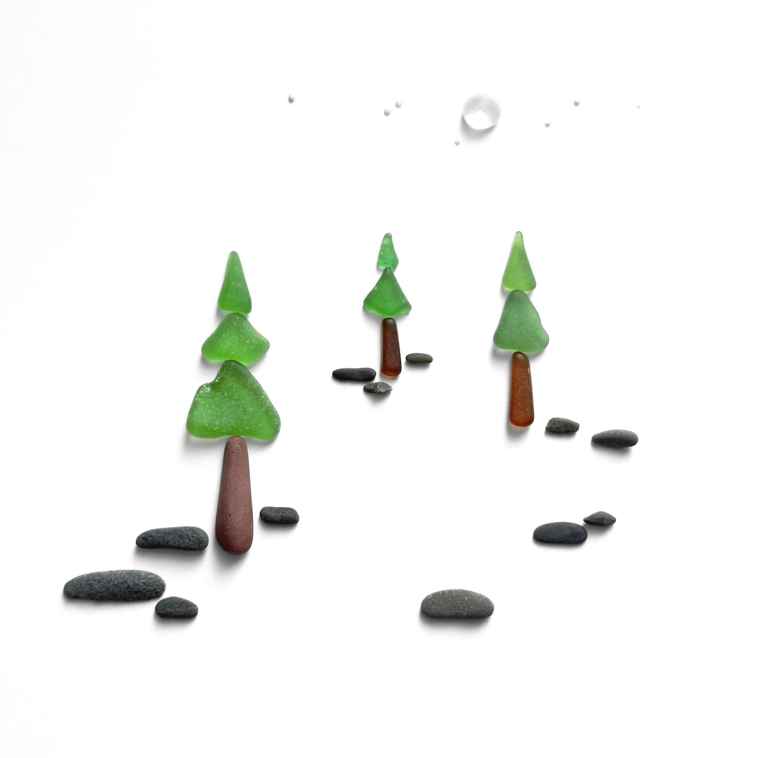 Three miniature evergreen trees stand beneath a glowing moon, with a trail of smooth beach stones winding through the clearing.