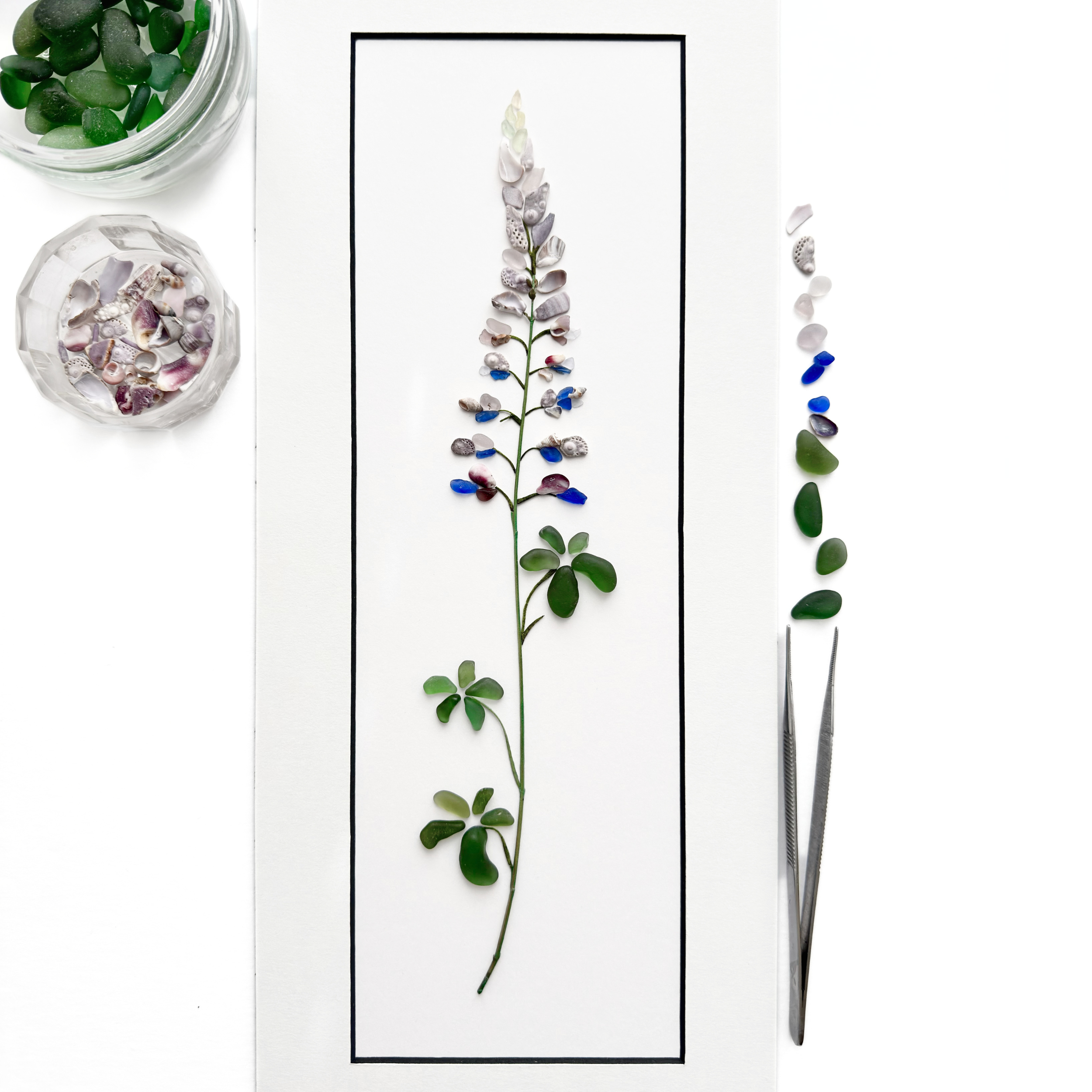 Studio process view of a sea glass lupine with sorted shells, glass pieces, and tweezers.