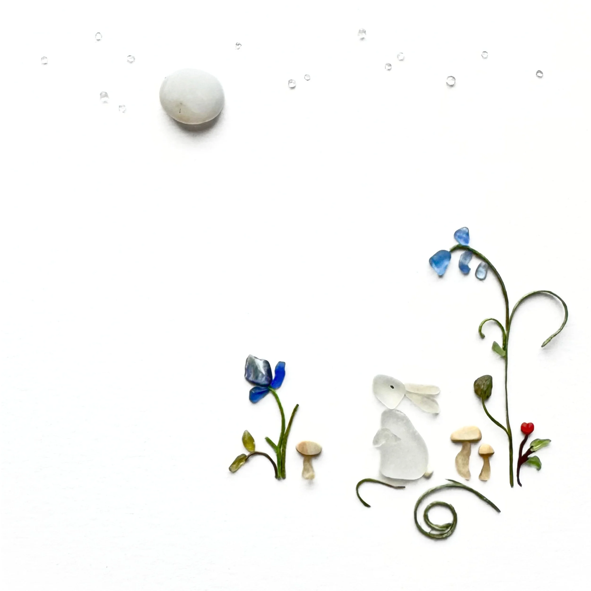 Original sea glass artwork featuring a small white rabbit sitting among blue flowers, miniature mushrooms, and curling botanical stems beneath a round pale beach stone moon against a white background.