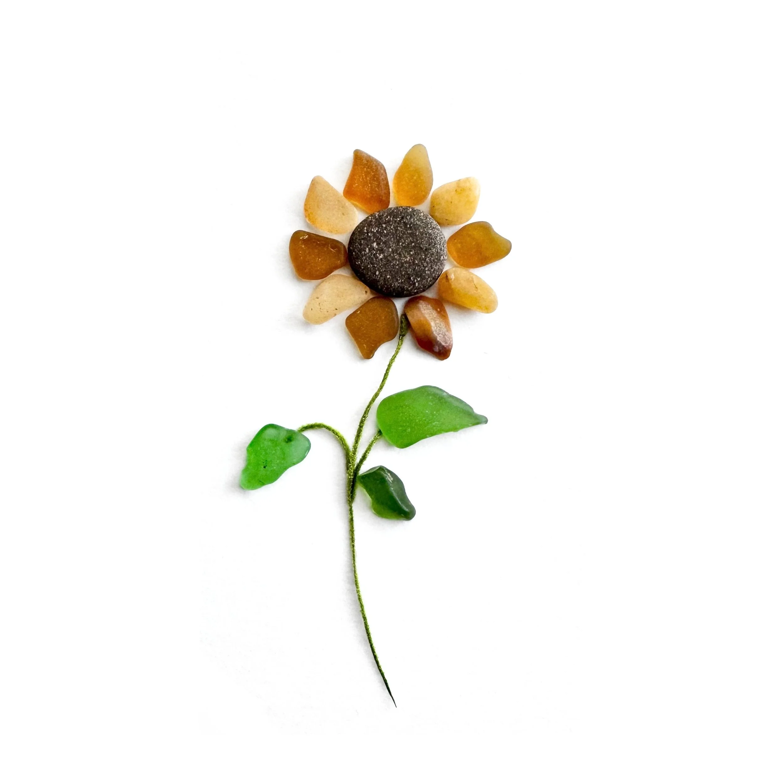 Original sunflower artwork featuring golden petals of sea glass, beach agate, and shell fragments, a dark beach stone center, green sea glass leaves, and a delicate botanical stem on white background.