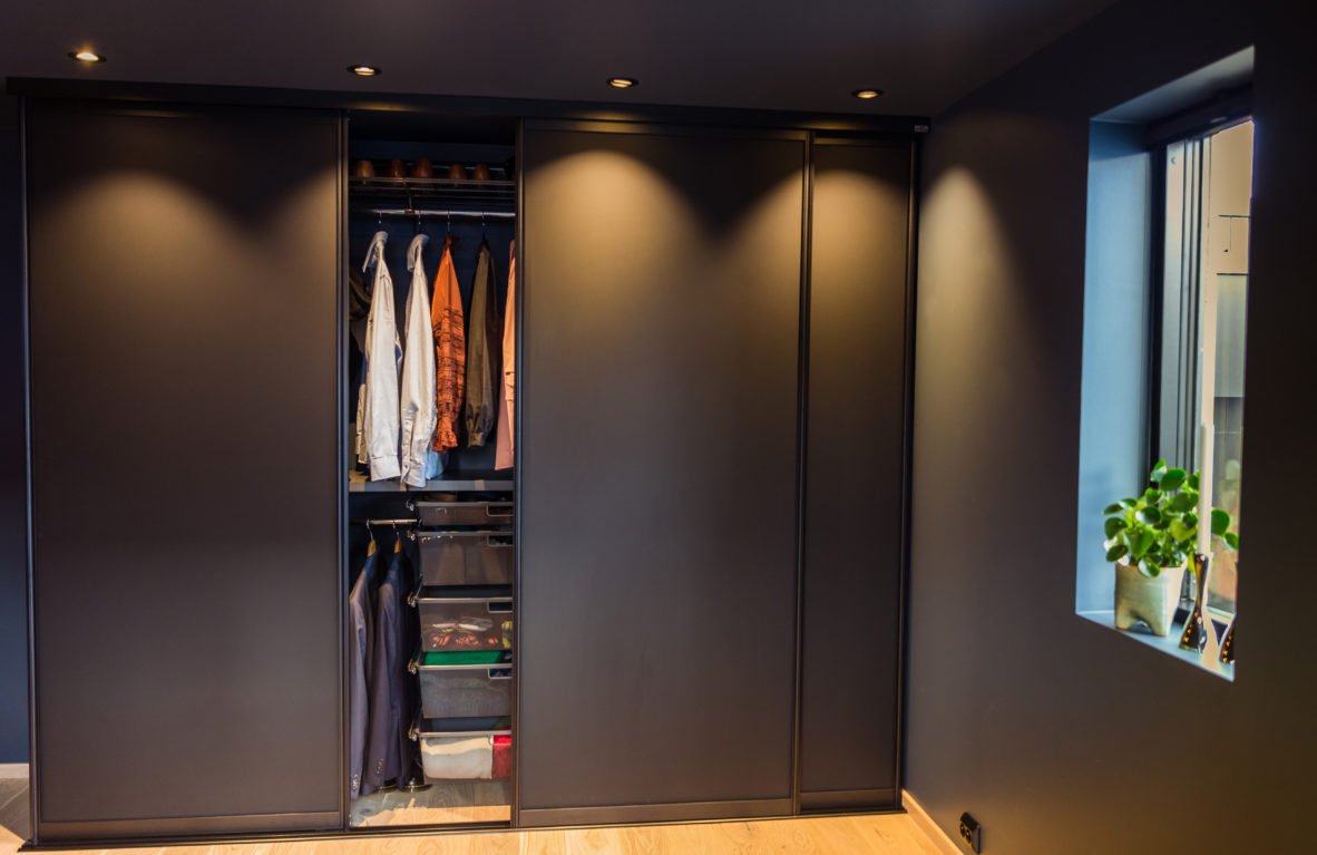 A dark sliding wardrobe with some clothes hanging and storage boxes inside, next to a window with a potted plant.