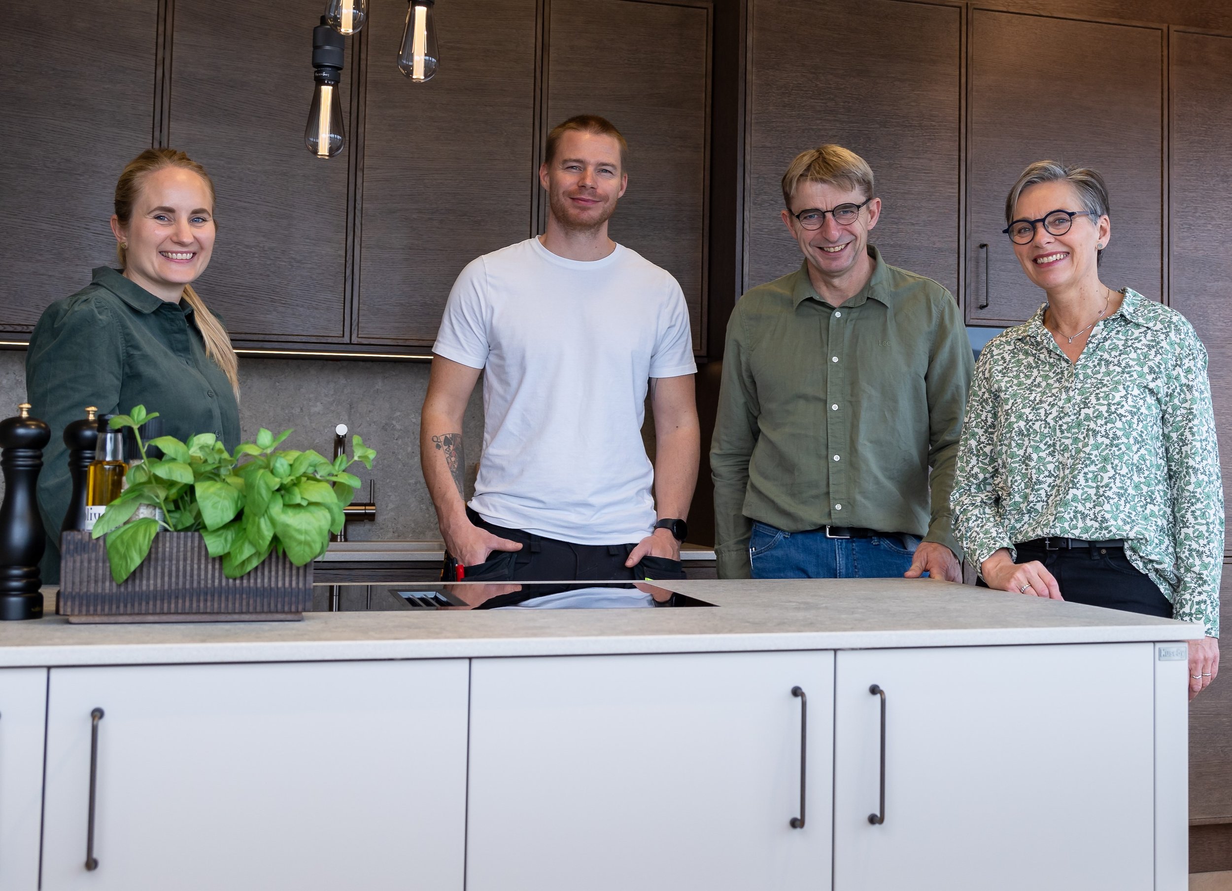 Fire personer står bak en kjøkkenøy, smilende, i et moderne kjøkken, med tre personer på høyre og en til venstre. Kjøkkenet har mørke skap og moderne lys.