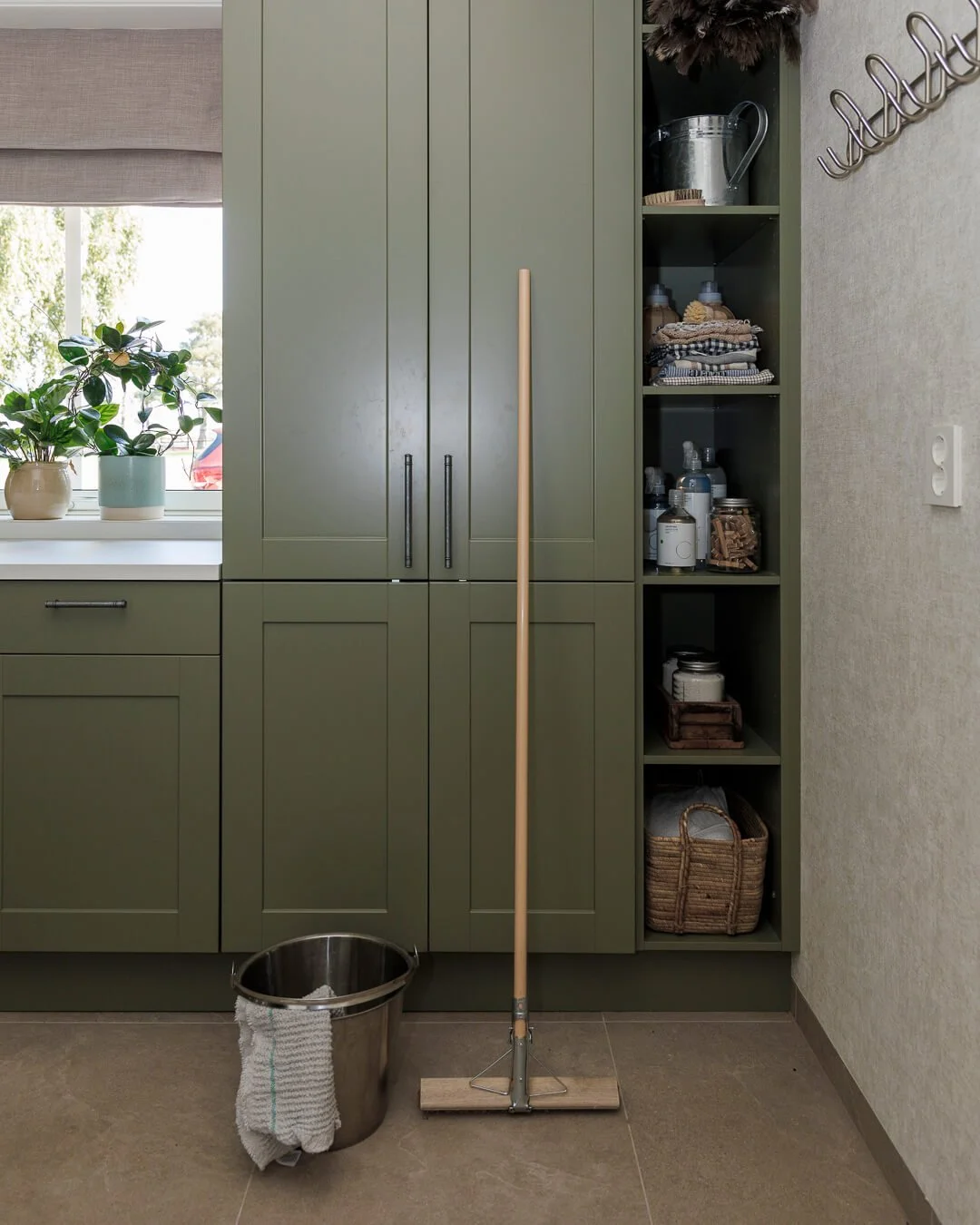 Kjøkken med grønne skap, vindu med blomsterpotter, beisebørste, mugge, oppbevaringshyller med vaskemidler, tøykluter, glasskrukker, kurv, og en strykejern.