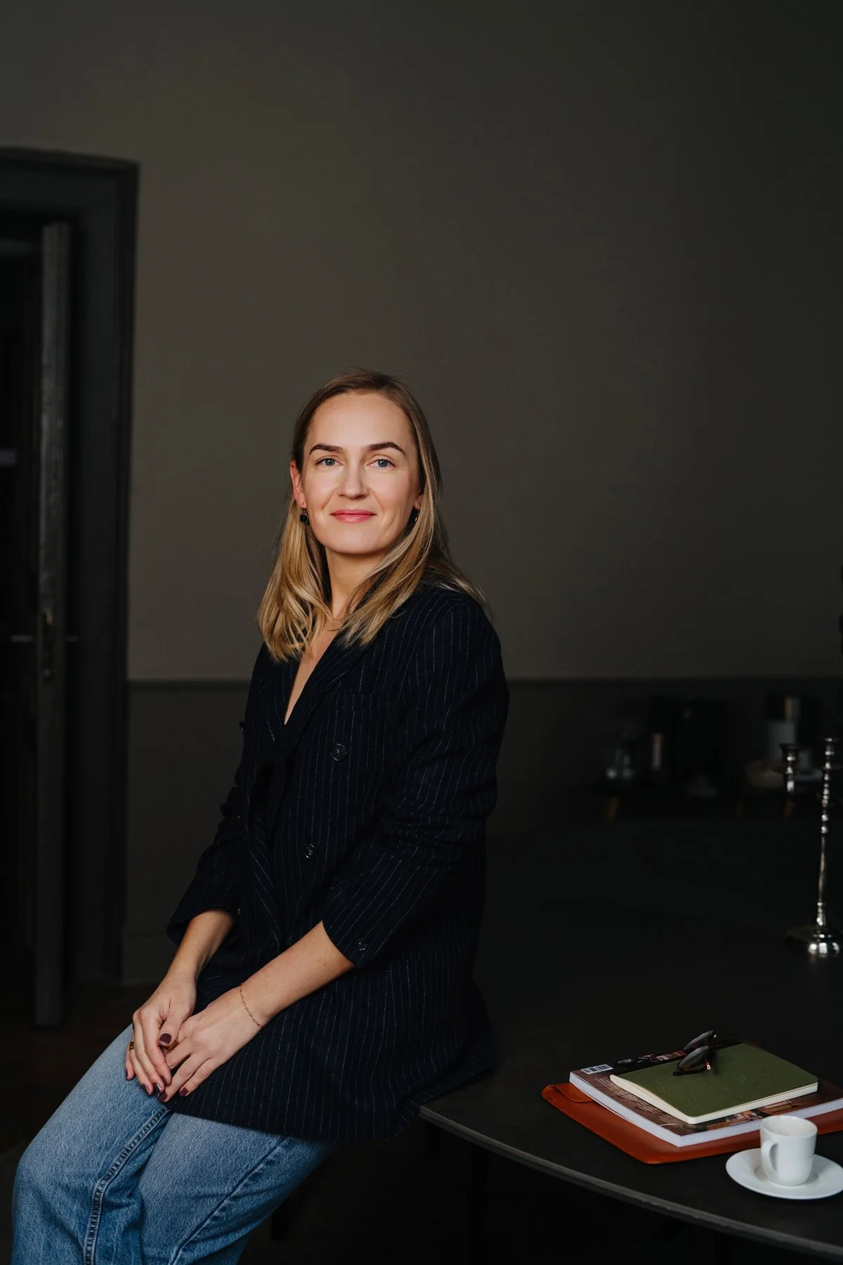 A woman with blonde hair wearing a black blazer with thin pinstripes and blue jeans, sitting on a table in a dimly lit room, with a notebook, glasses, and a cup on the table.