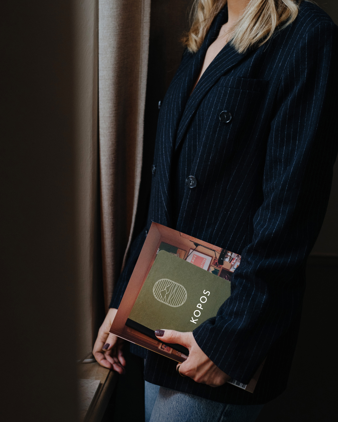 A woman wearing a dark pinstripe blazer holding a box with the logo 'KOPOS' on it, standing beside a window with curtains.