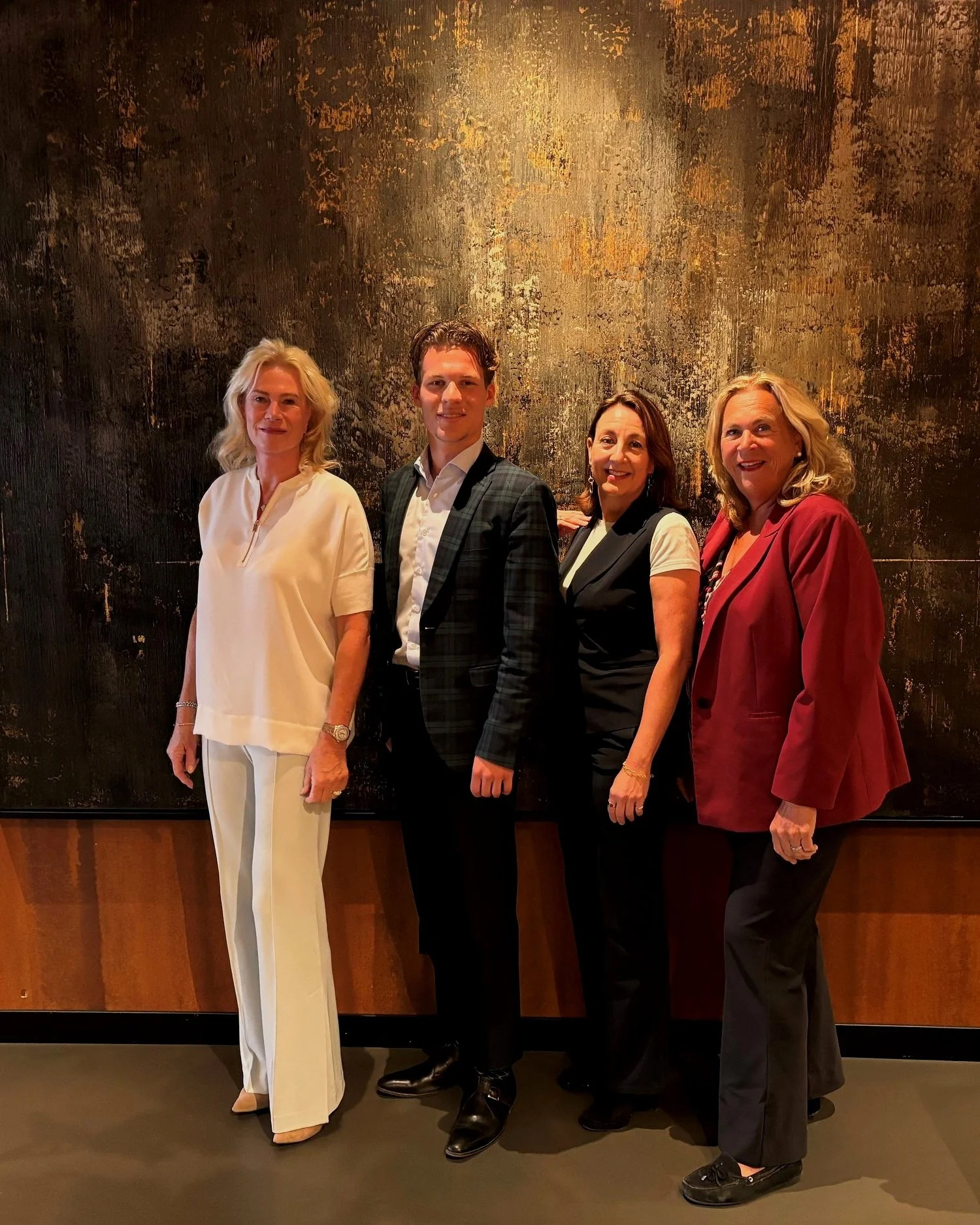 Vier mensen staan naast elkaar voor een abstract kunstwerk op een muur, gekleed in formele en semi-formele kleding.