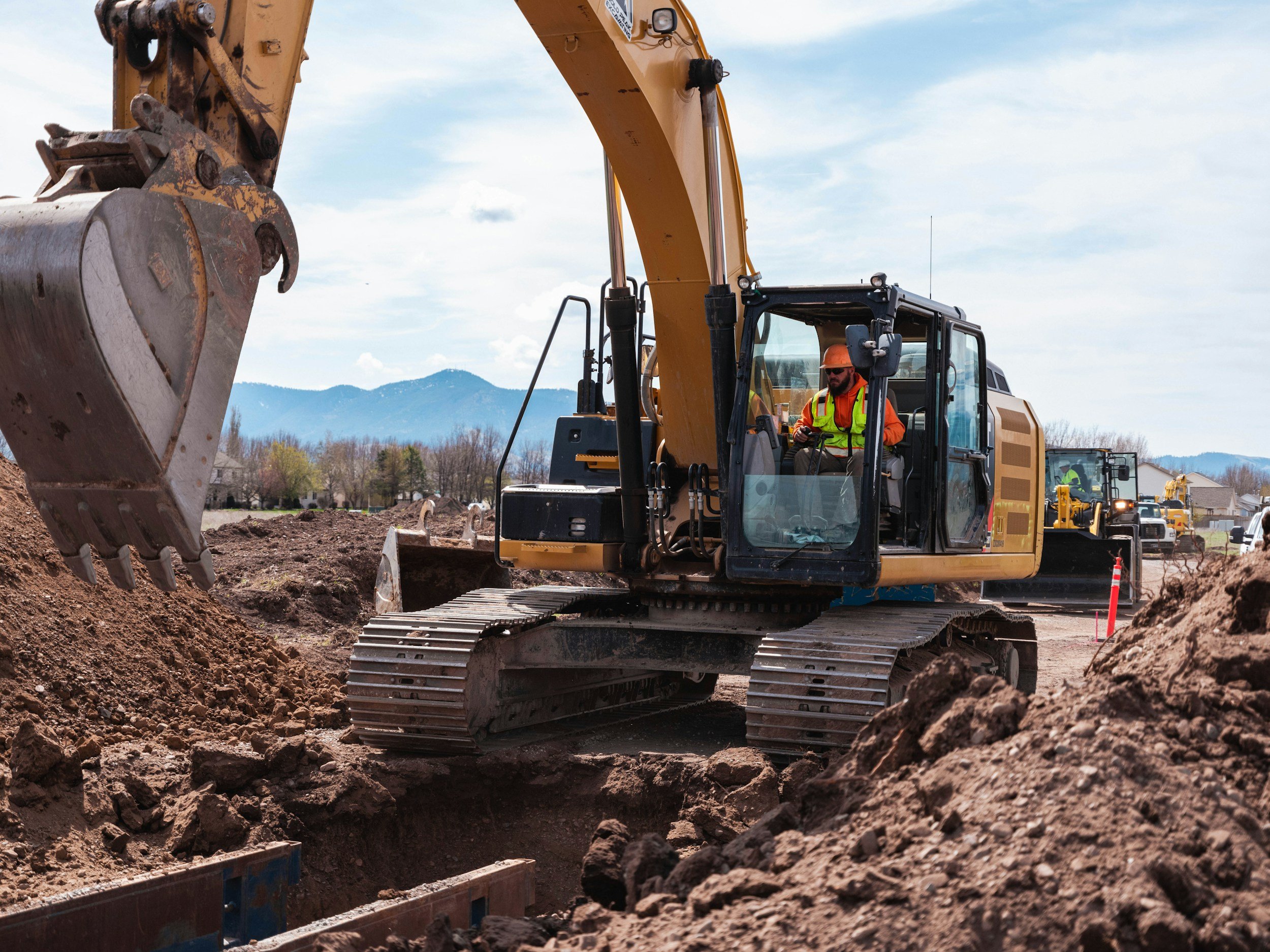 A construction worker operating a yellow excavator at a construction site during daytime, with a backdrop of mountains and a cloudy sky.