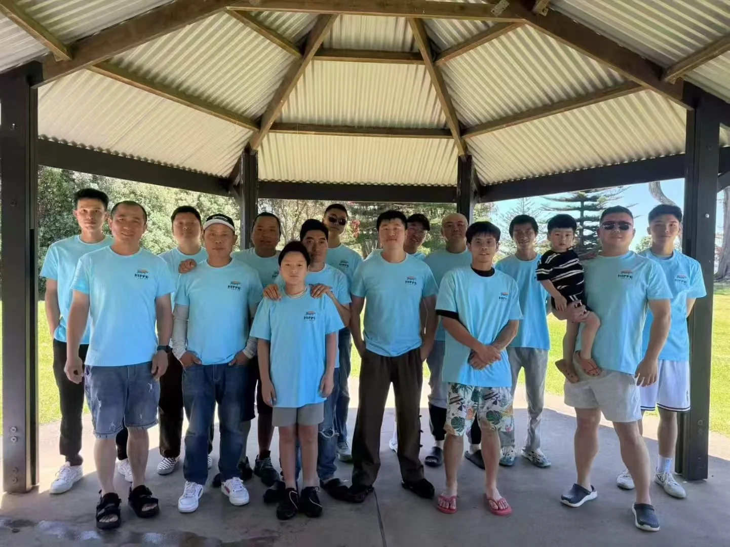 Group of fifteen people, mostly men, standing under a pavilion outdoors. They are wearing light blue t-shirts, and some are wearing sunglasses. One child is being held by a man. The background shows greenery and trees.