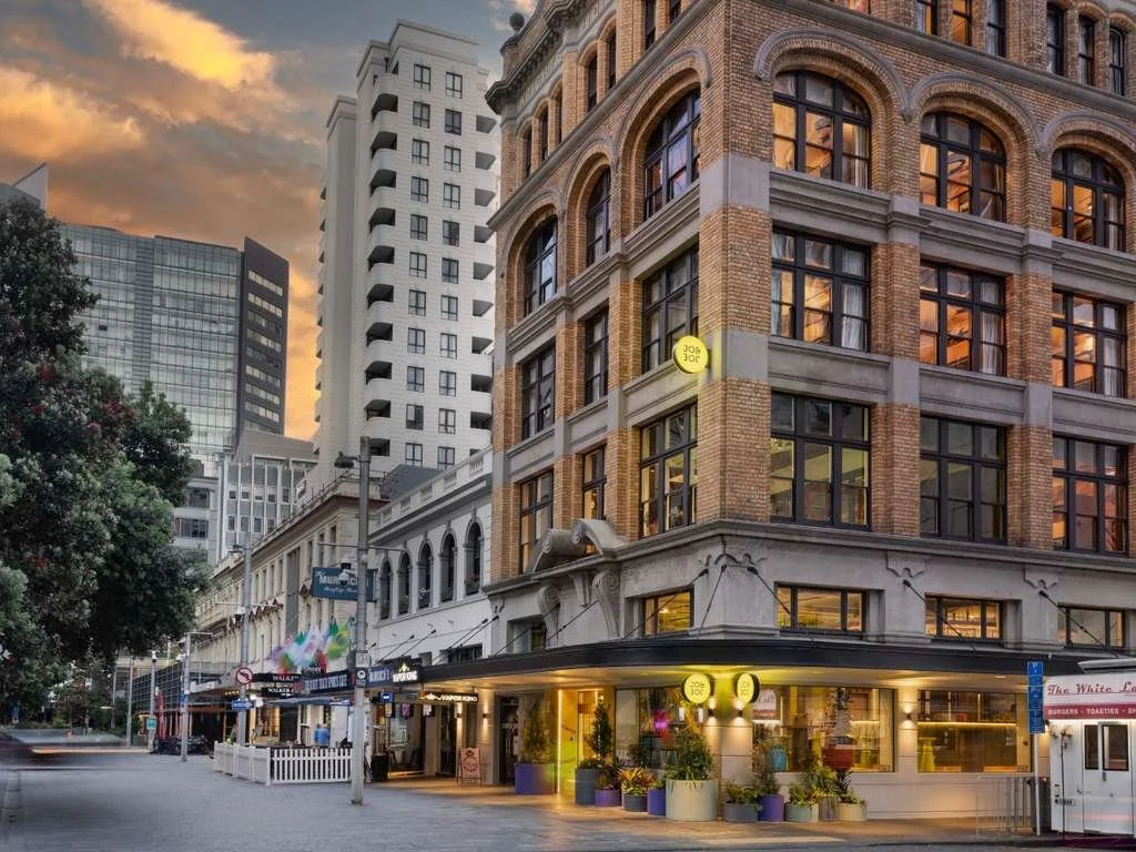 City street scene during sunset with tall buildings, trees, and a corner building with a sign that reads "Job", storefronts, and outdoors seating areas.