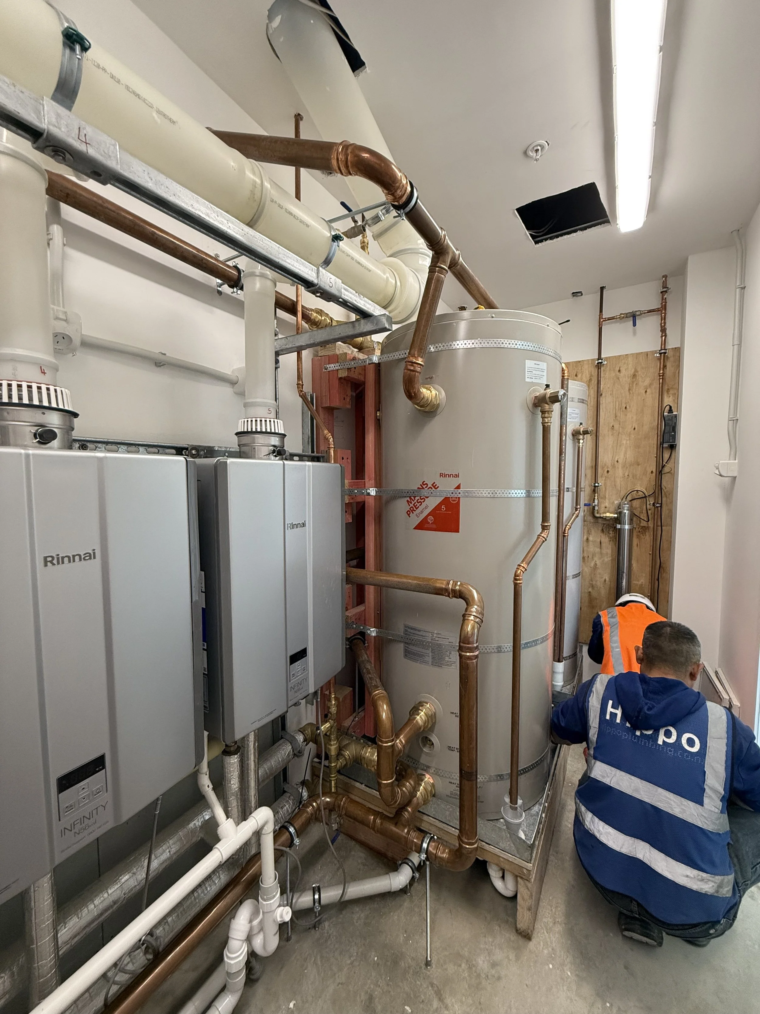 A construction worker kneeling on the floor installing or inspecting plumbing pipes in a utility room with white walls and a gray floor. There are multiple pipes, including copper and white PVC, and two gray Rinnai Infinity tankless water heaters mounted on the wall.