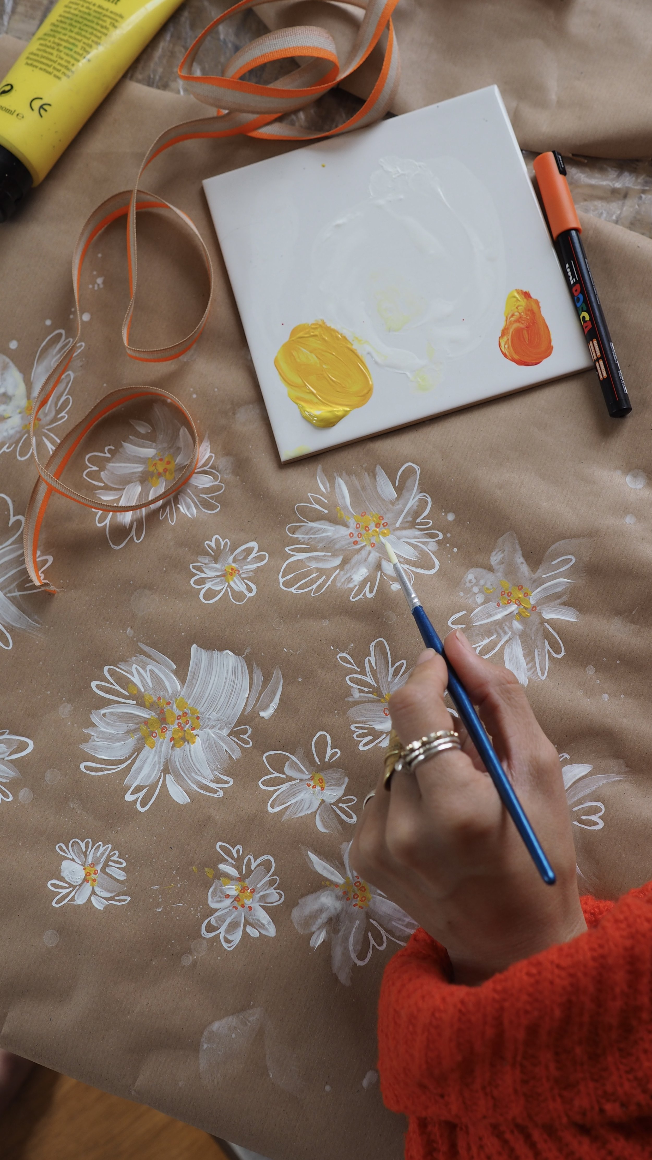 A hand painting white flowers on brown paper with yellow and orange paint. There is a paintbrush, a tube of yellow paint, and a piece of ribbon nearby. A paint marker is also visible.