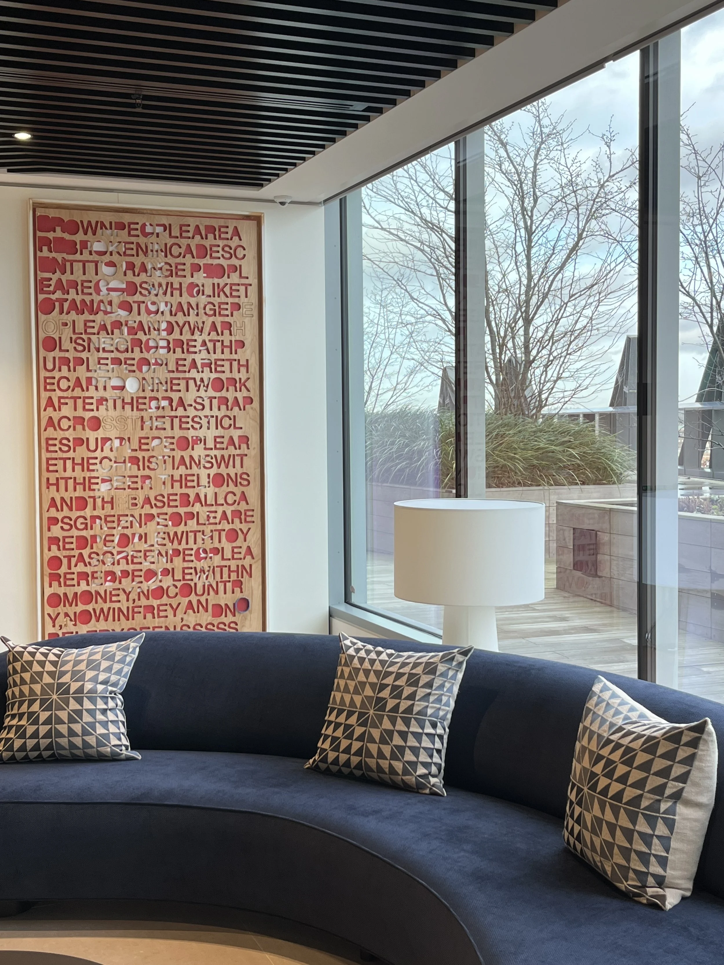 A modern lounge area with a curved navy blue sofa adorned with patterned pillows, next to large floor-to-ceiling windows revealing a barren tree and cloudy sky outside. A white cylindrical lampshade sits on a small side table, and a decorative wooden panel with red cut-out letters is on the wall.